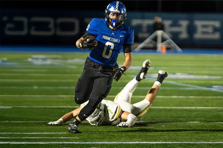 A football player from the University of Minnesota running with the ball while a player from the opposing team lies on the ground behind him on the football field.