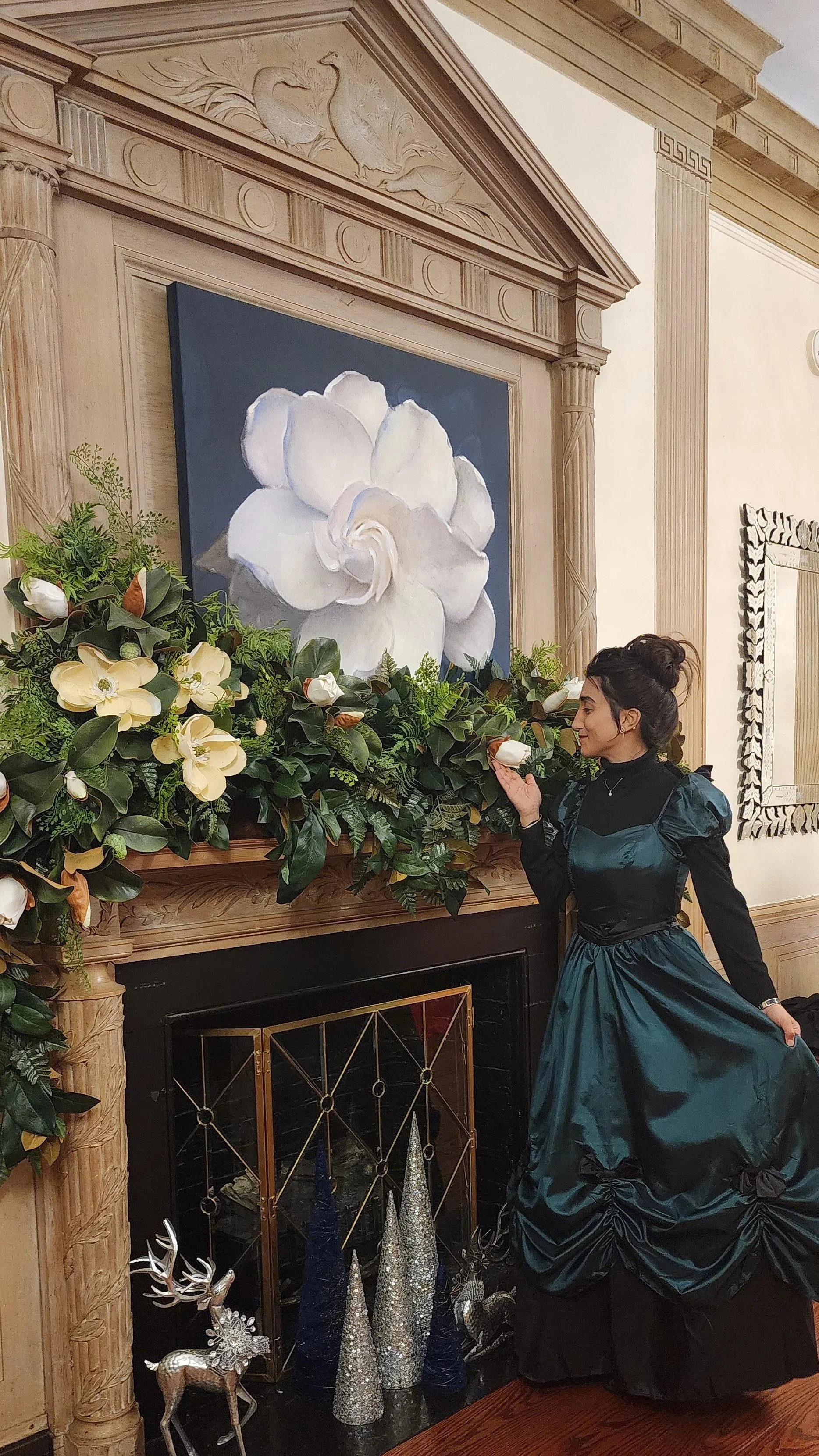 Woman in teal evening gown smelling a white flower near a decorated fireplace with a floral artwork on the wall above.