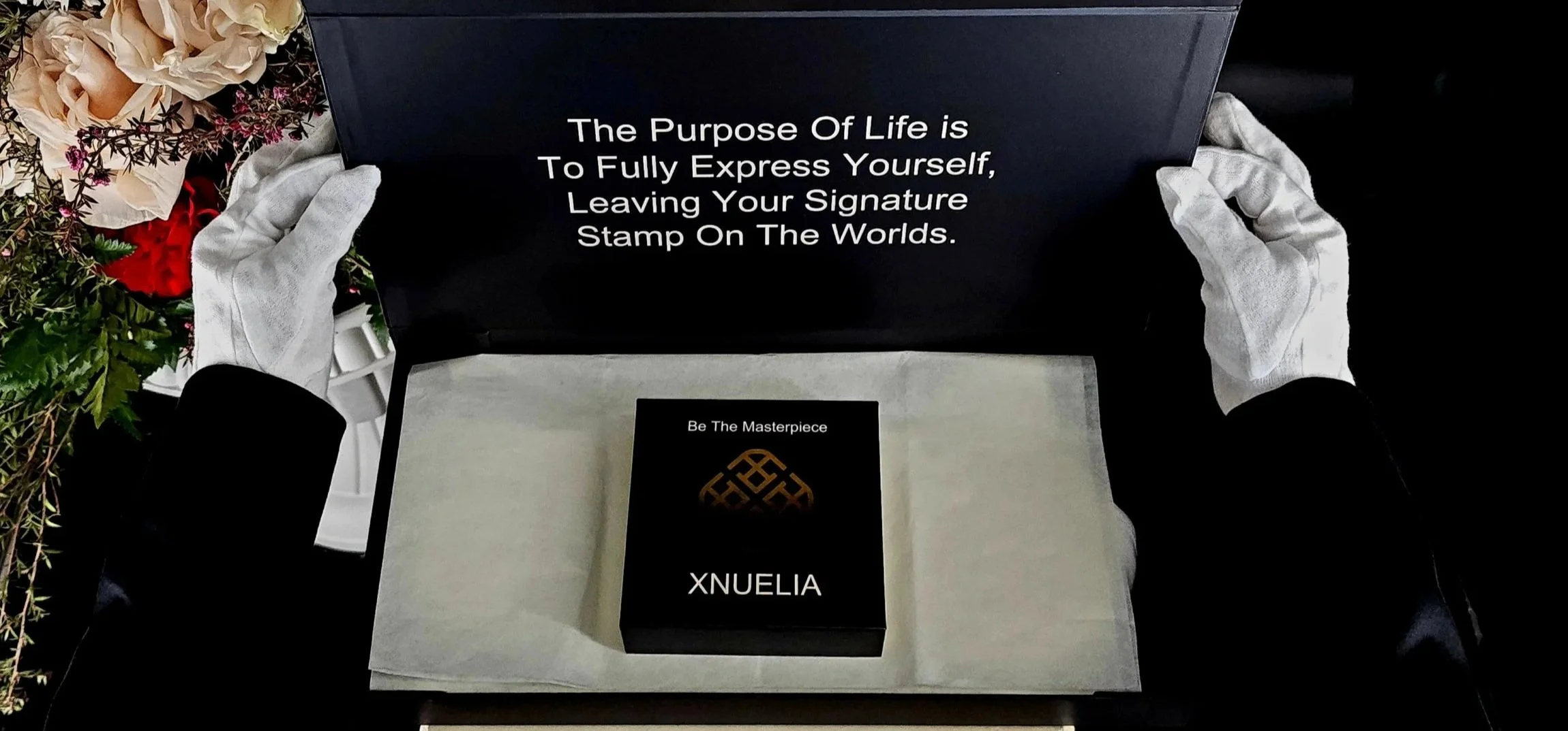 A person wearing white gloves holding an open black box with a motivational quote inside. The quote reads, "The Purpose of Life is To Fully Express Yourself, Leaving Your Signature Stamp on The Worlds." Inside the box, there is a smaller black package with a gold logo and the text "Be The Masterpiece" and "XNUELIA." There are flowers on the side of the scene.