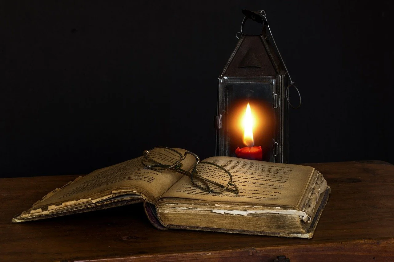 An old, open book with glasses resting on it on a wooden surface, illuminated by a candle inside a lantern in the background.