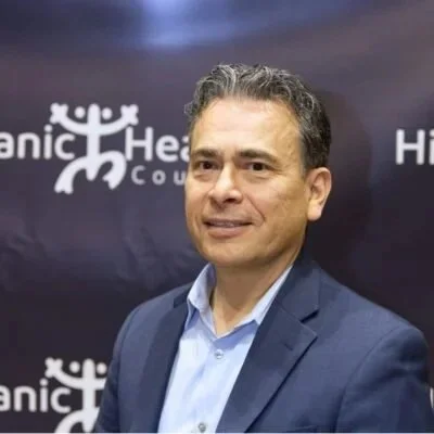 A man in a blue suit and light blue shirt standing in front of a dark background with the logo of the American Heart Association.