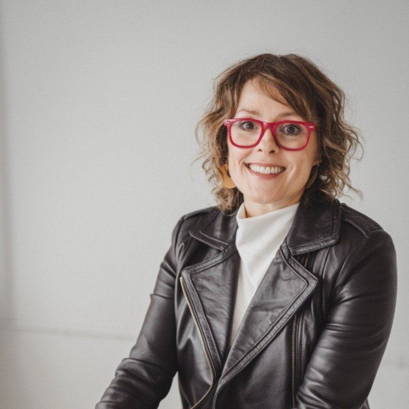 A woman with curly hair, red glasses, and a smile, wearing a black leather jacket and a white top, standing against a plain background.