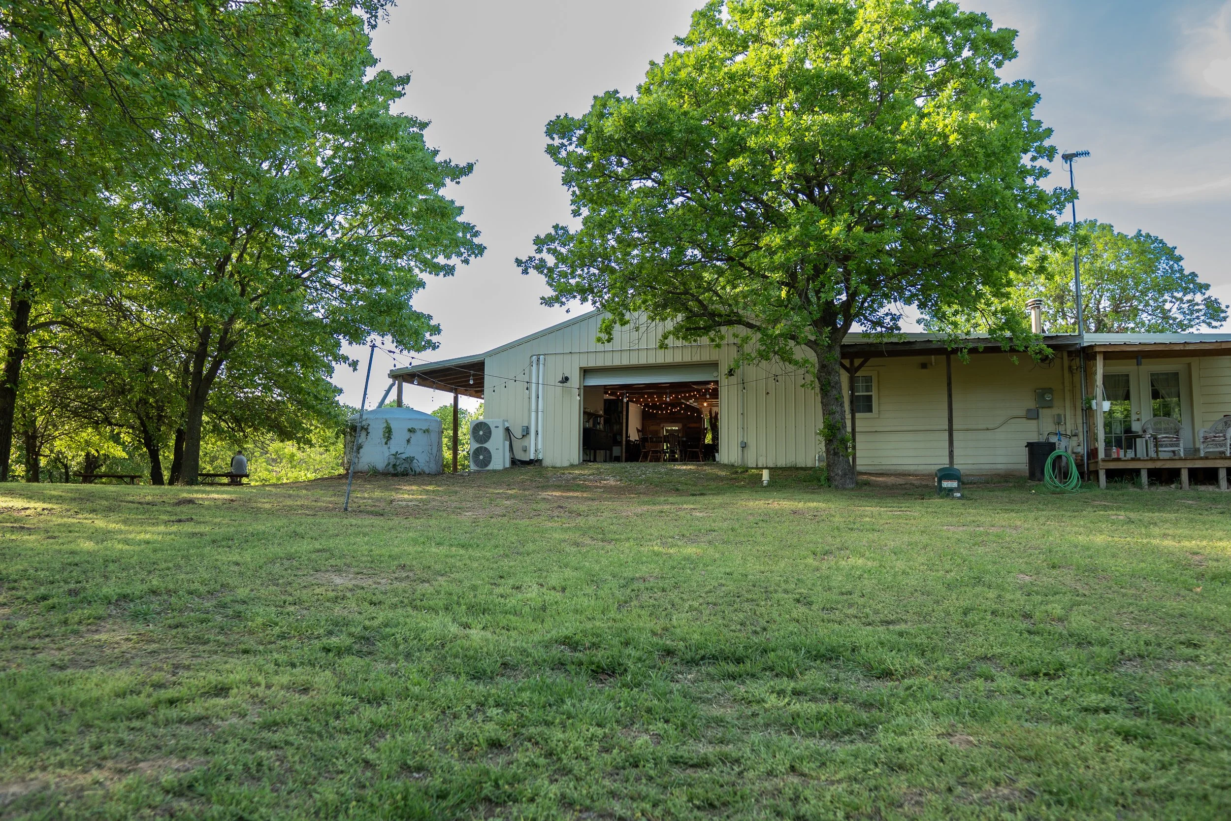 Green backyard with large trees and a barn with open garage revealing string lights inside, surrounding a grassy area and outdoor seating.