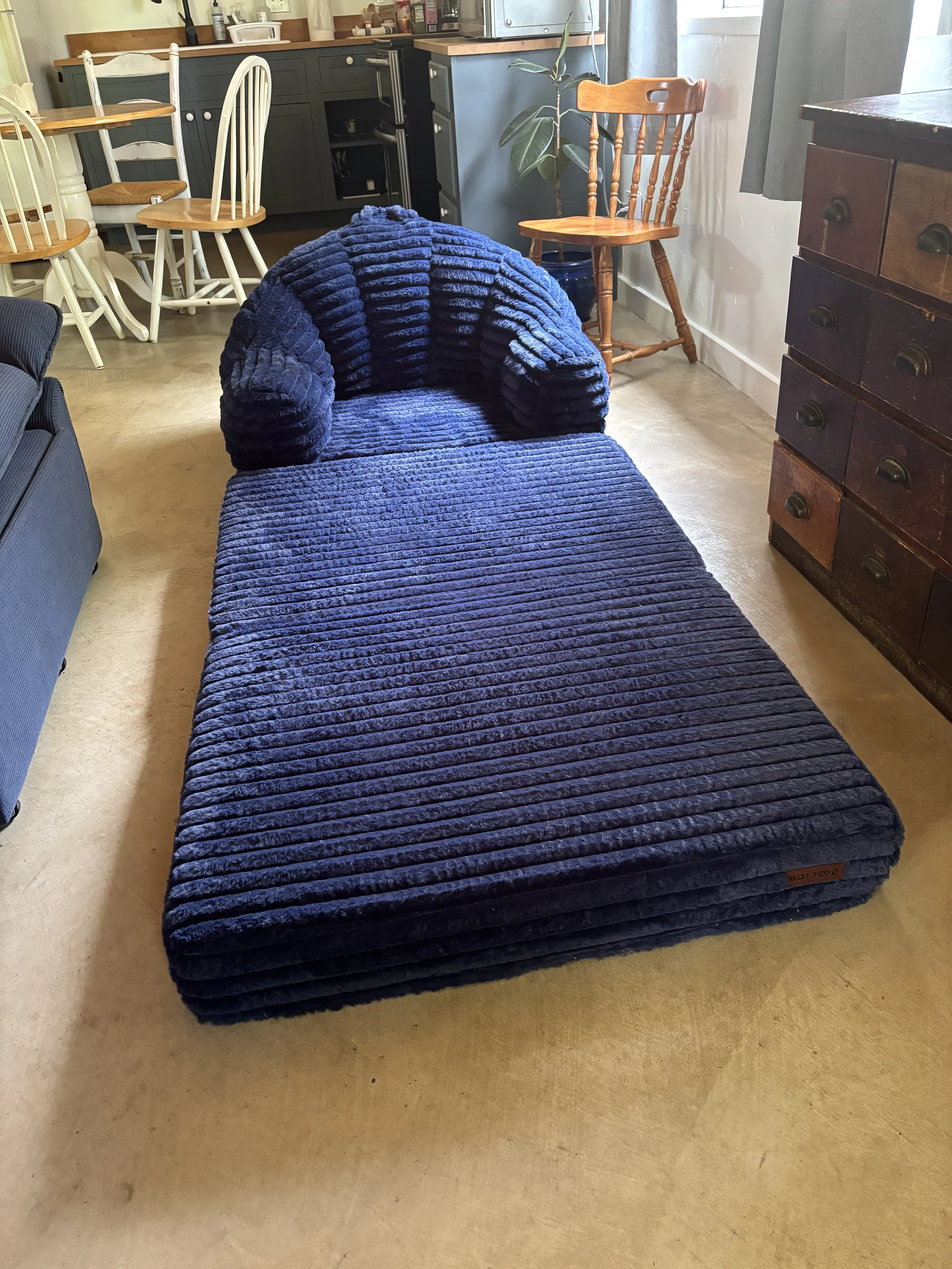 A dark blue, plush, nubby upholstered lounge chair with a matching ottoman in a room with wooden furniture, including a wooden chest of drawers and dining chairs, and a small plant near a window with gray curtains.