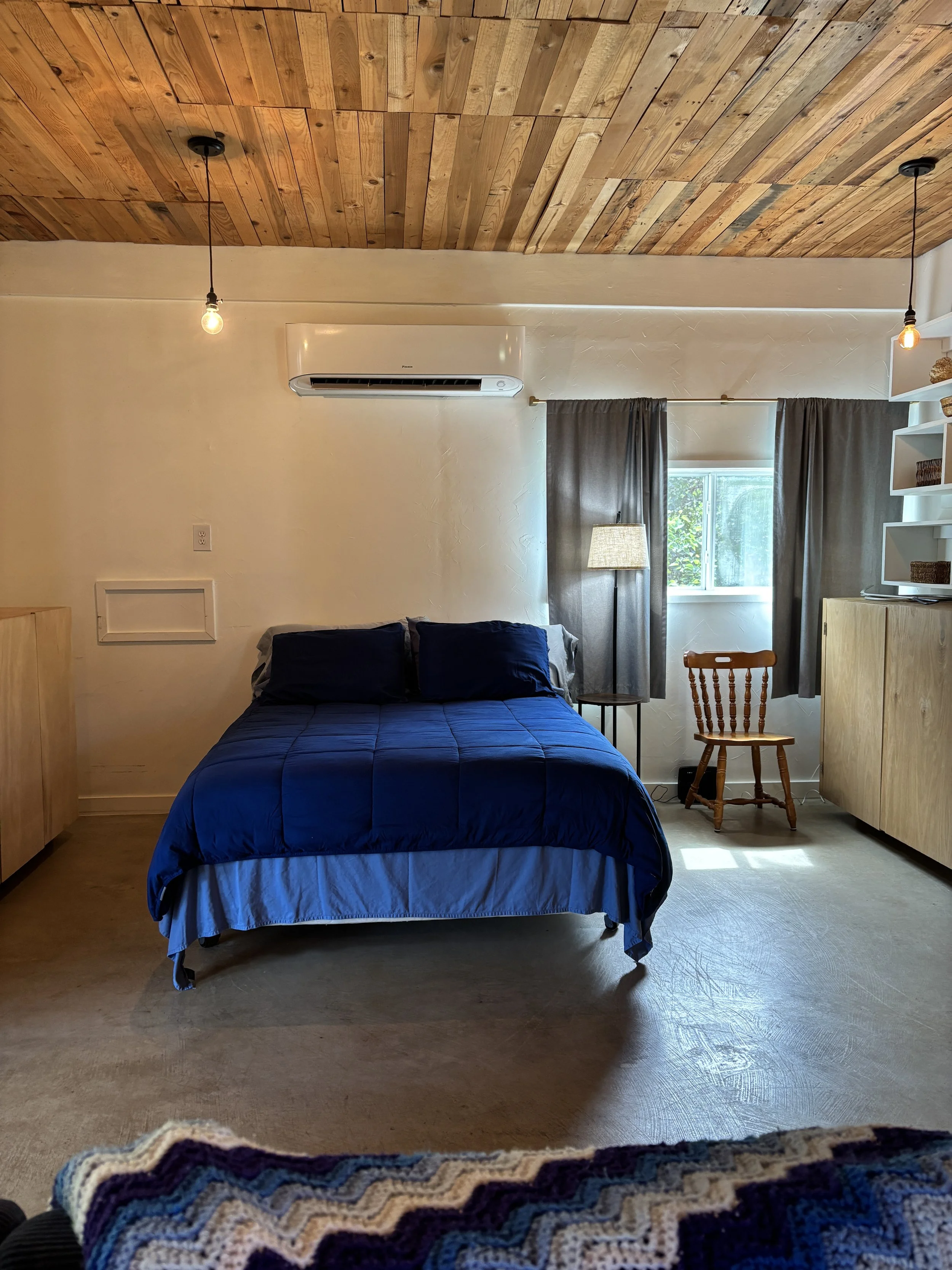 Interior of a bedroom with a bed dressed in a blue comforter and matching pillows. The ceiling is made of wood planks, and there are two hanging light bulbs. To the right, a window with gray curtains is partially open, letting in natural light. There