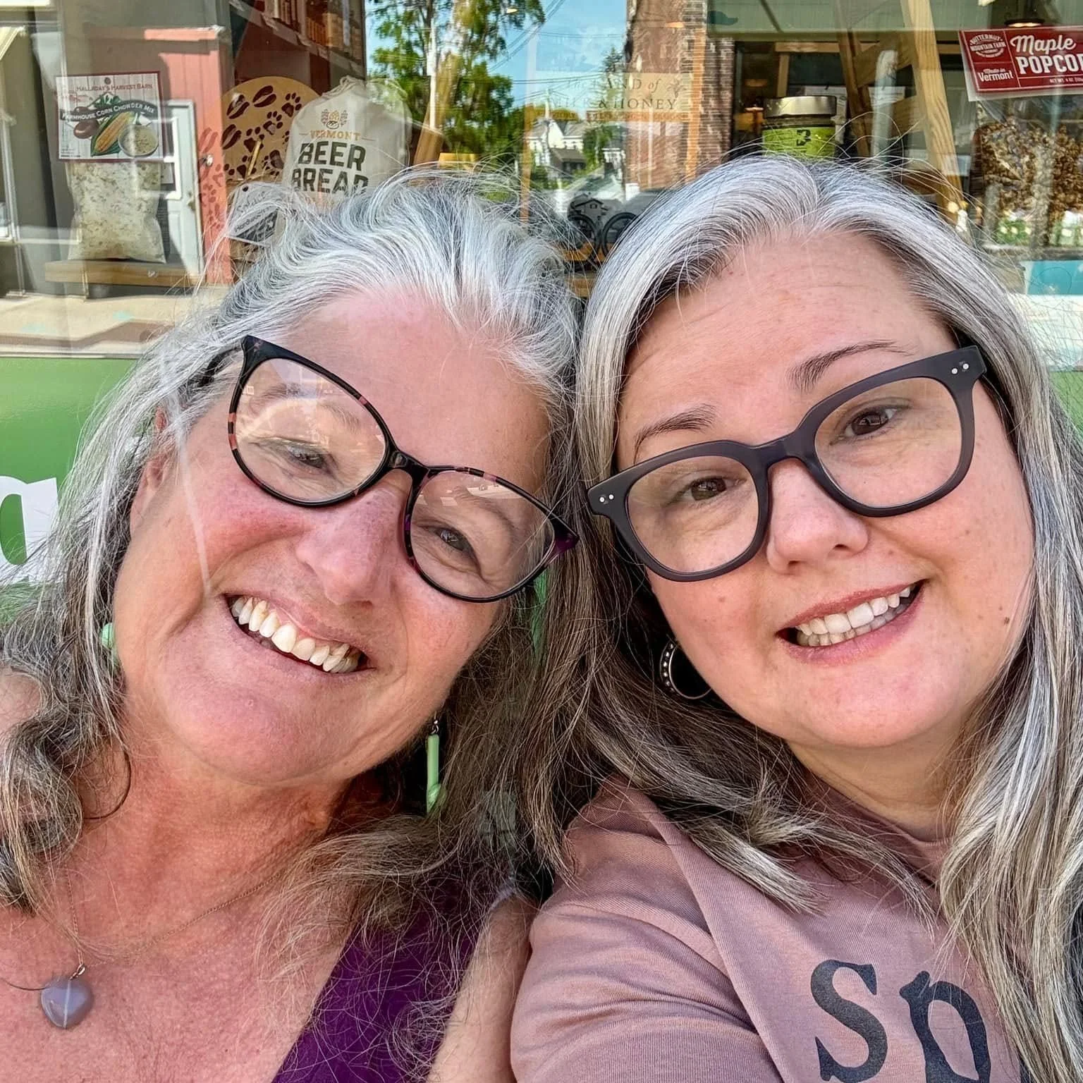 Two women smiling and wearing glasses, taking a selfie outside a storefront with signs and items in the background.