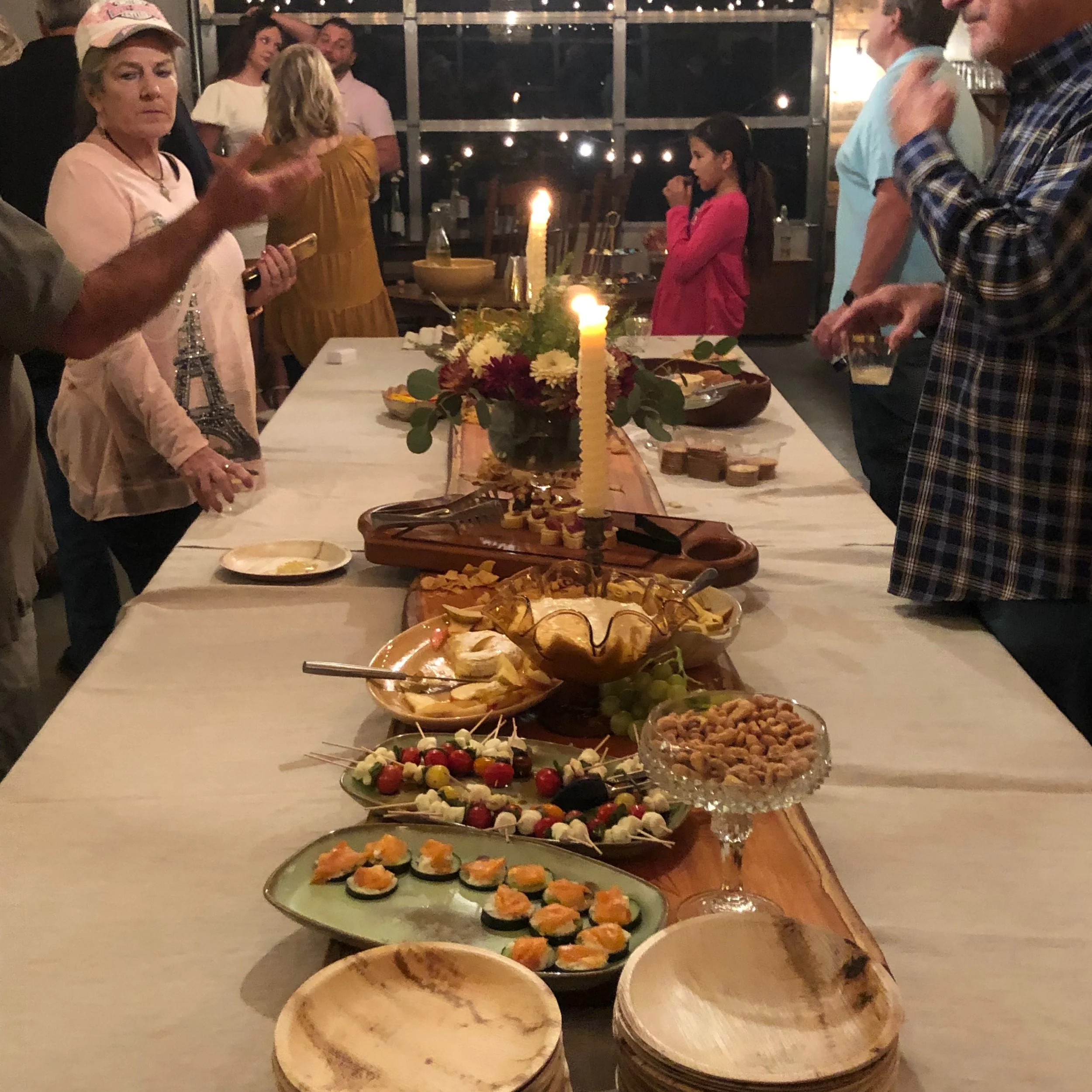 A buffet table with an assortment of appetizers and snacks, including sushi, cherry tomatoes with mozzarella and basil, and cheese spreads. In the background, people are gathered around, engaging in conversation, at what appears to be a social or cel