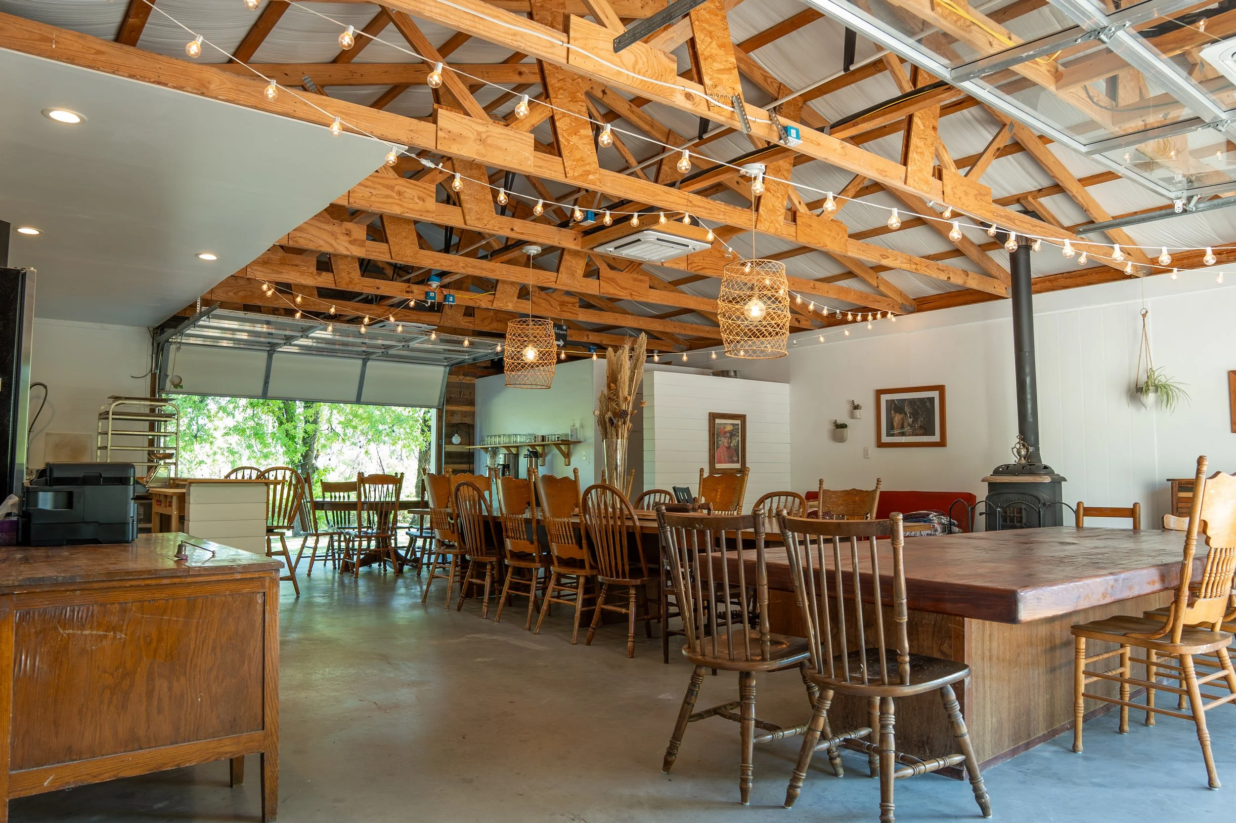 Interior of a cozy restaurant or cafe with a rustic design, featuring wooden tables, chairs, and exposed wooden ceiling beams decorated with string lights and hanging lanterns.