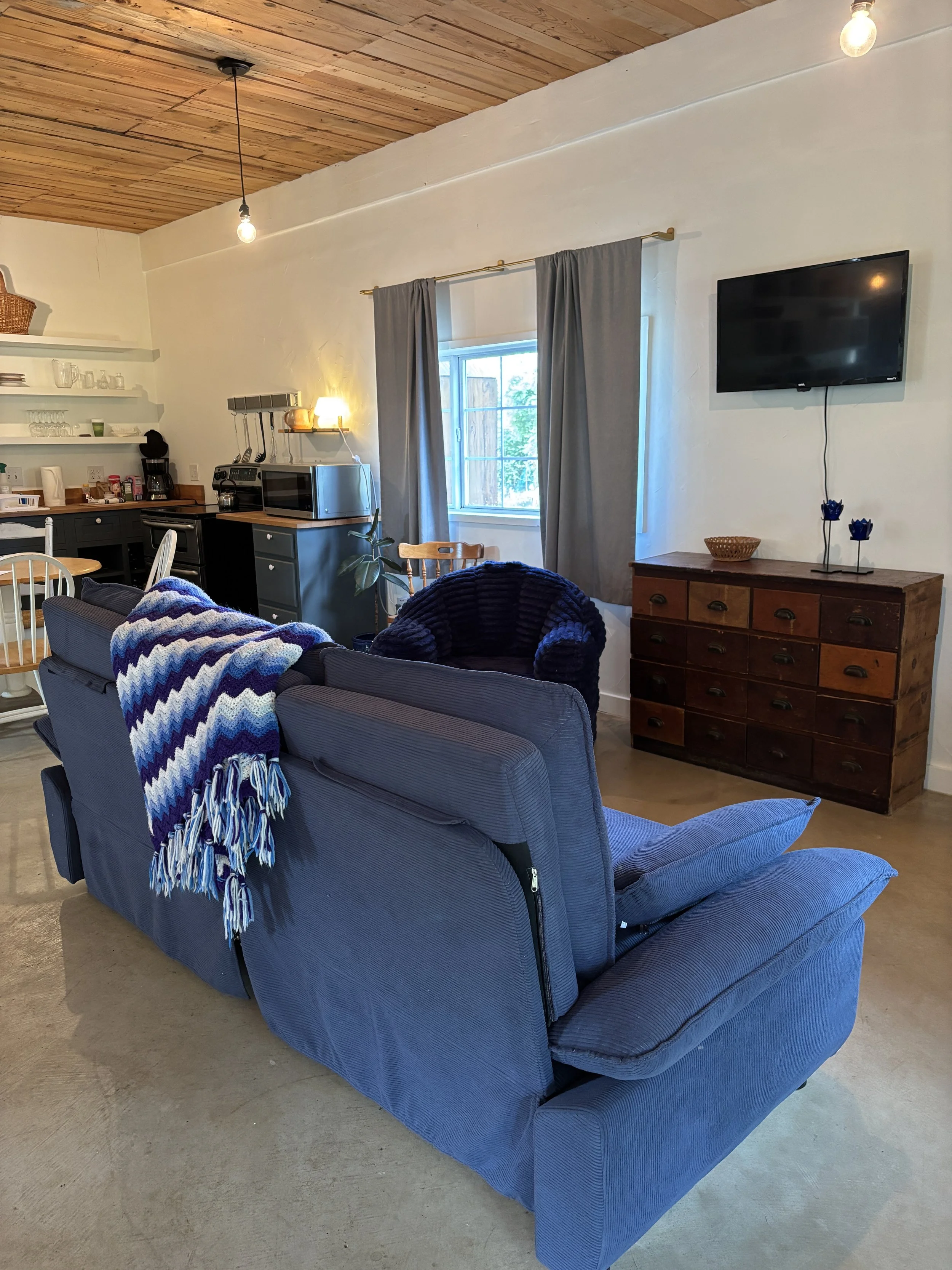 Living room with a blue sofa, a blanket draped over the back, and a wooden floor with a wooden ceiling. There is a window with gray curtains, a TV mounted on the wall, and a wooden dresser underneath the TV. The room has various appliances and decor,