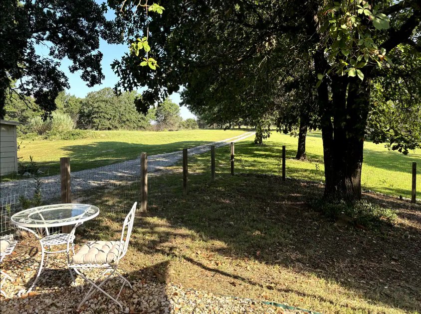 A backyard scene with a small round metal table and two matching chairs, one with a cushion, on a gravel patio. A wooden fence runs along the yard, which has green grass, a large tree providing shade, and a path leading into a field of open grass and