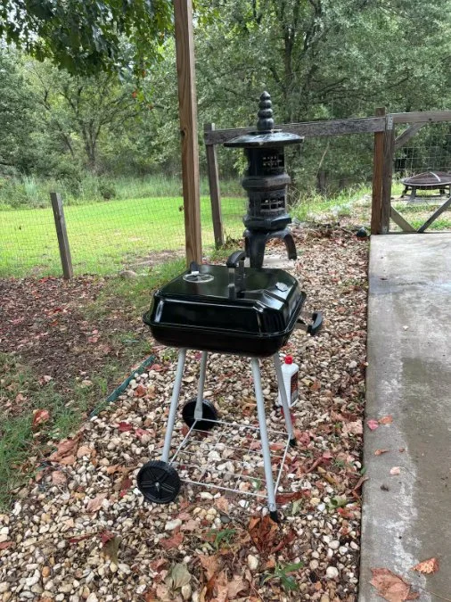 A black kettle grill on a small mobile stand with a metal rod, situated outdoors on a gravel surface, next to a concrete patio, with a wooden fence and trees in the background.