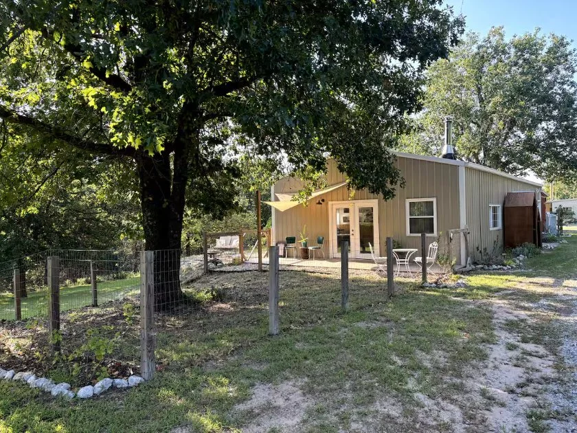 A house with a metal exterior, two windows, and a sliding glass door, surrounded by trees and a simple yard with a wooden fence, outdoor seating, and a dirt pathway.
