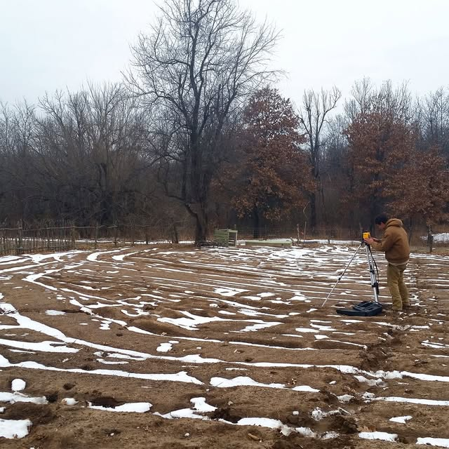 Kelly using surveying equipment in muddy field with patches of snow.