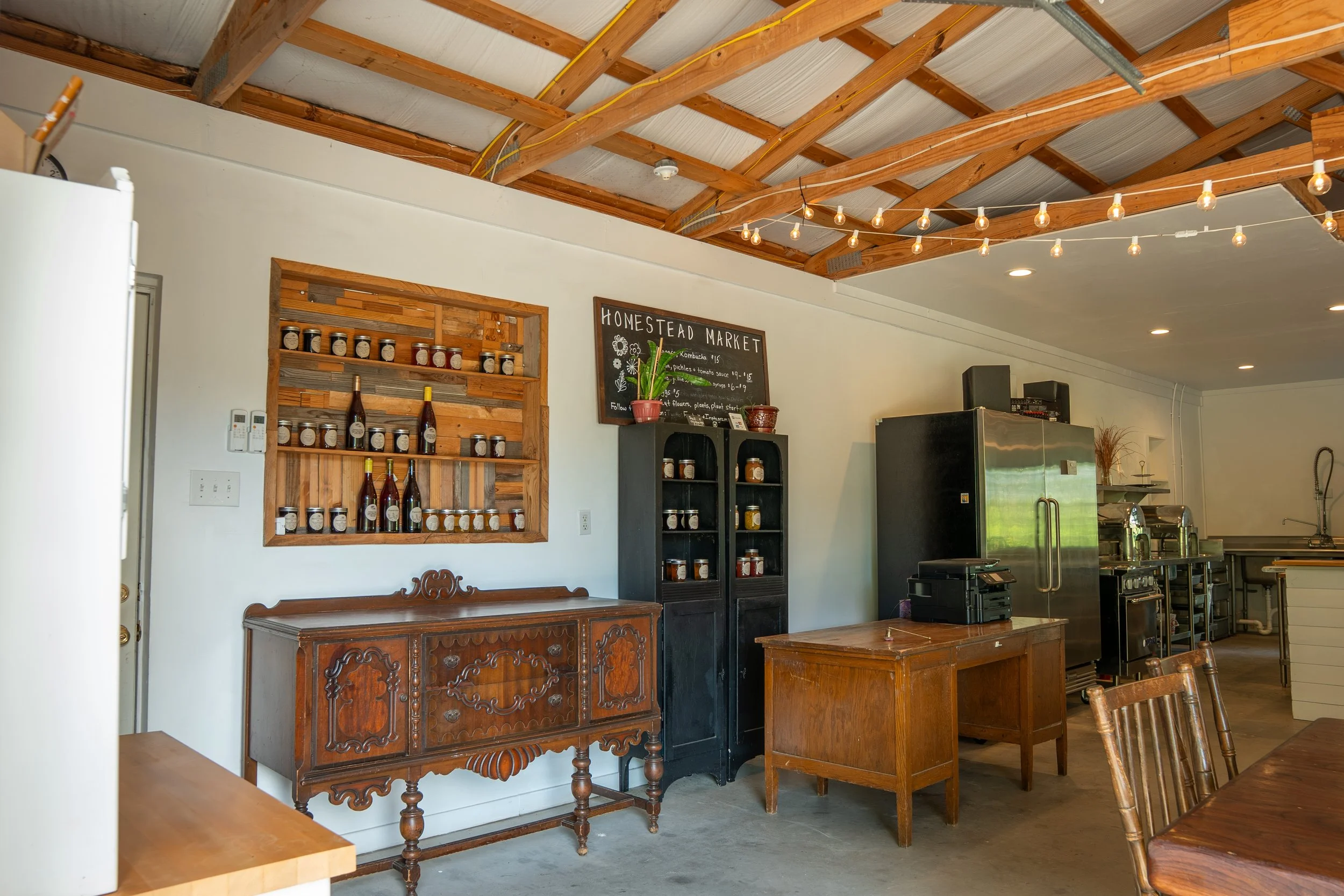 Interior of a rustic market or cafe with wooden ceiling beams, string lights, shelves with jars and bottles, a blackboard sign, vintage furniture, and industrial kitchen equipment in the background.