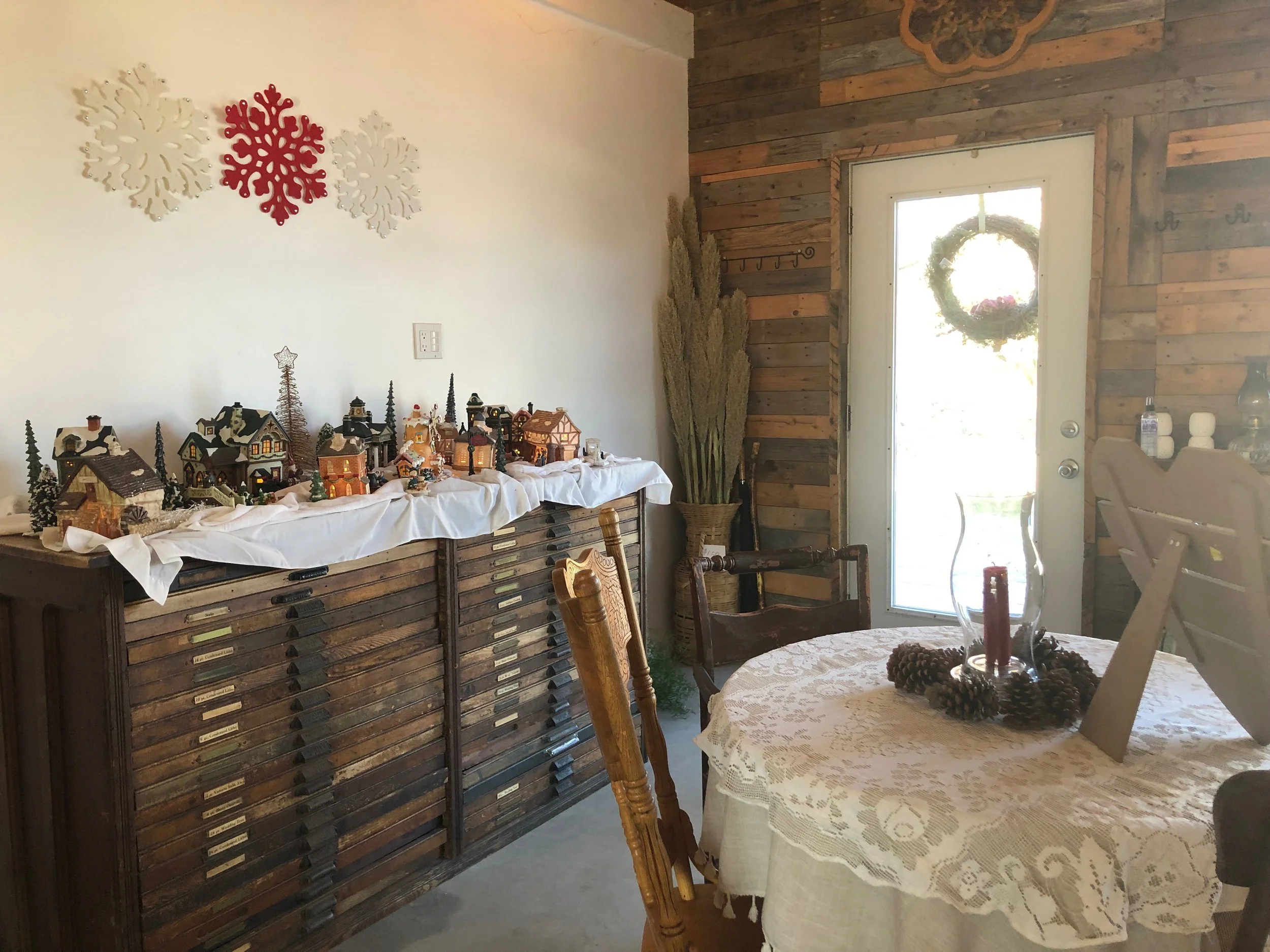 A room decorated for Christmas with a table displaying miniature houses and Christmas decorations. The wall on the left has white and red snowflake ornaments. There is a wooden wall and a door with a wreath outside. A chandelier with pinecones hangs 