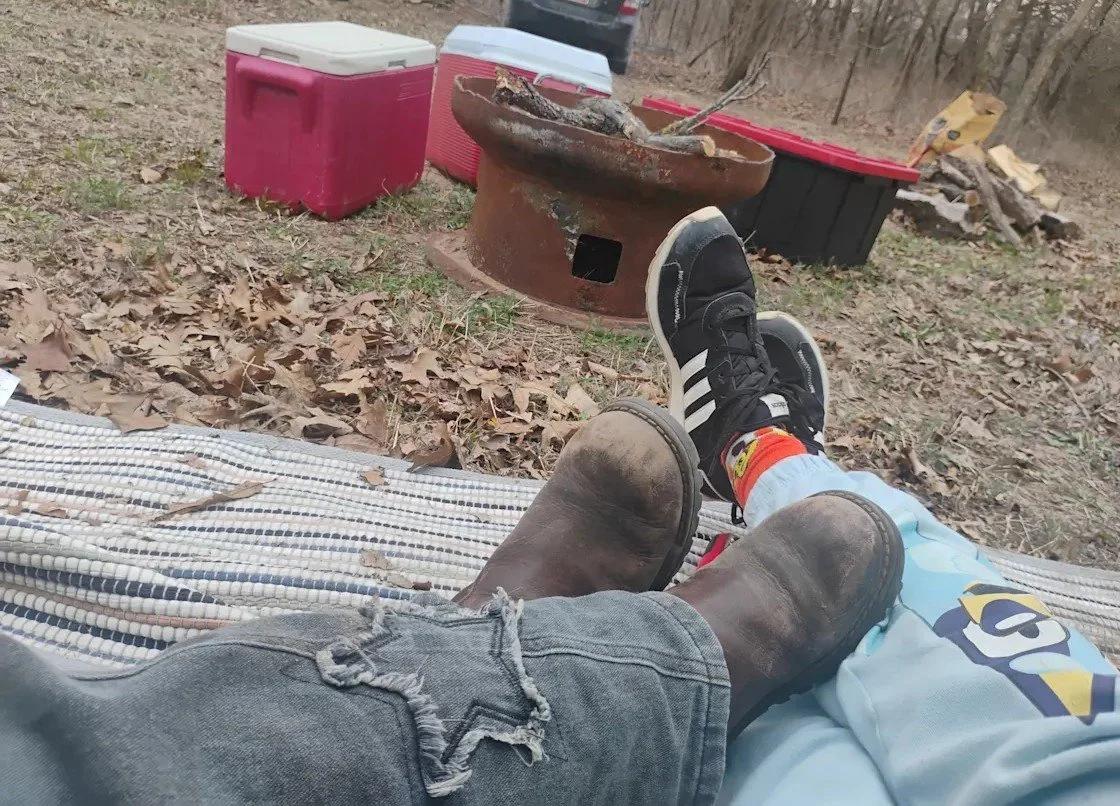 People relaxing outdoors in a wooded area, with their legs and feet visible, one person wearing black sneakers and the other wearing cowboy boots, while looking at a fire pit and coolers in the background.