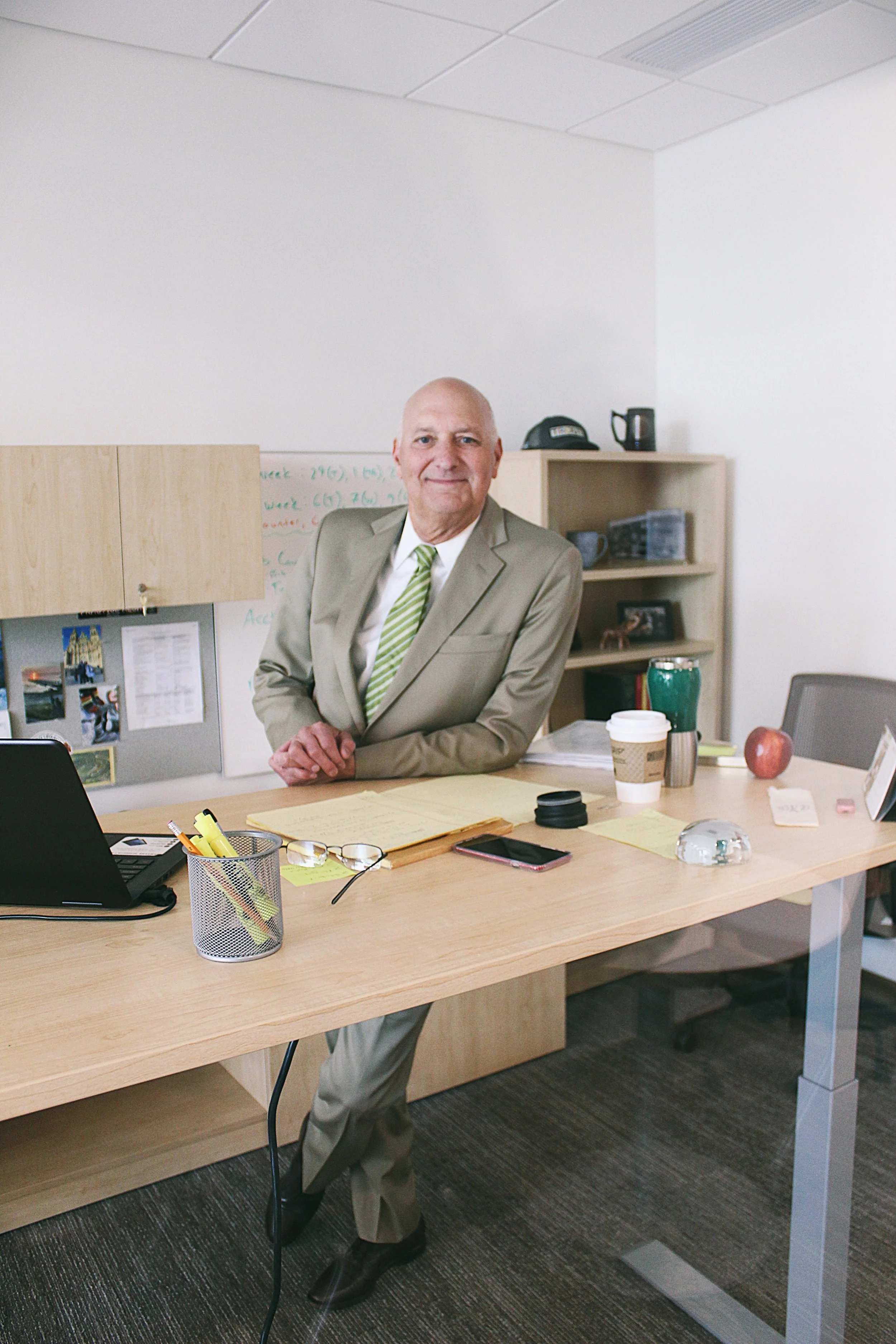 A man in a beige suit with a green striped tie standing behind an office desk, surrounded by papers, coffee cups, and office supplies in a modern office with white walls and wooden furniture.