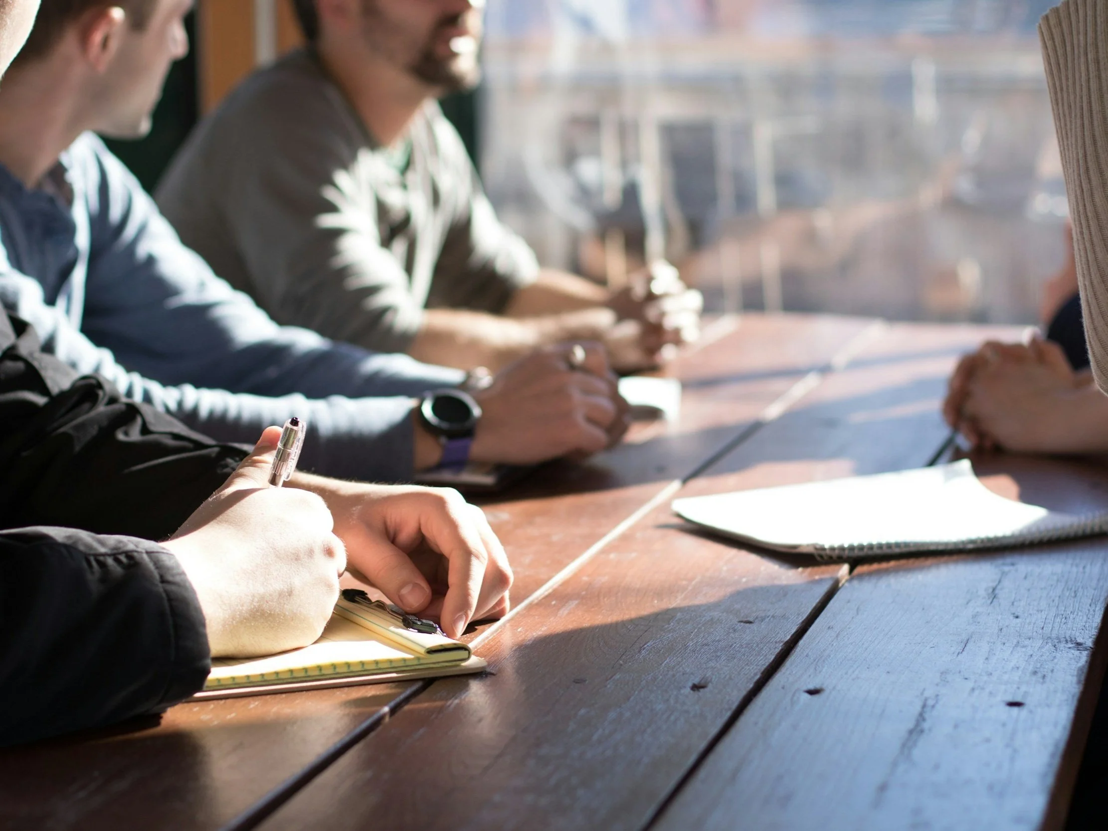 Four people sitting at a wooden table, engaged in a meeting or discussion, focusing on notes and documents. The image is taken in a well-lit indoor setting.