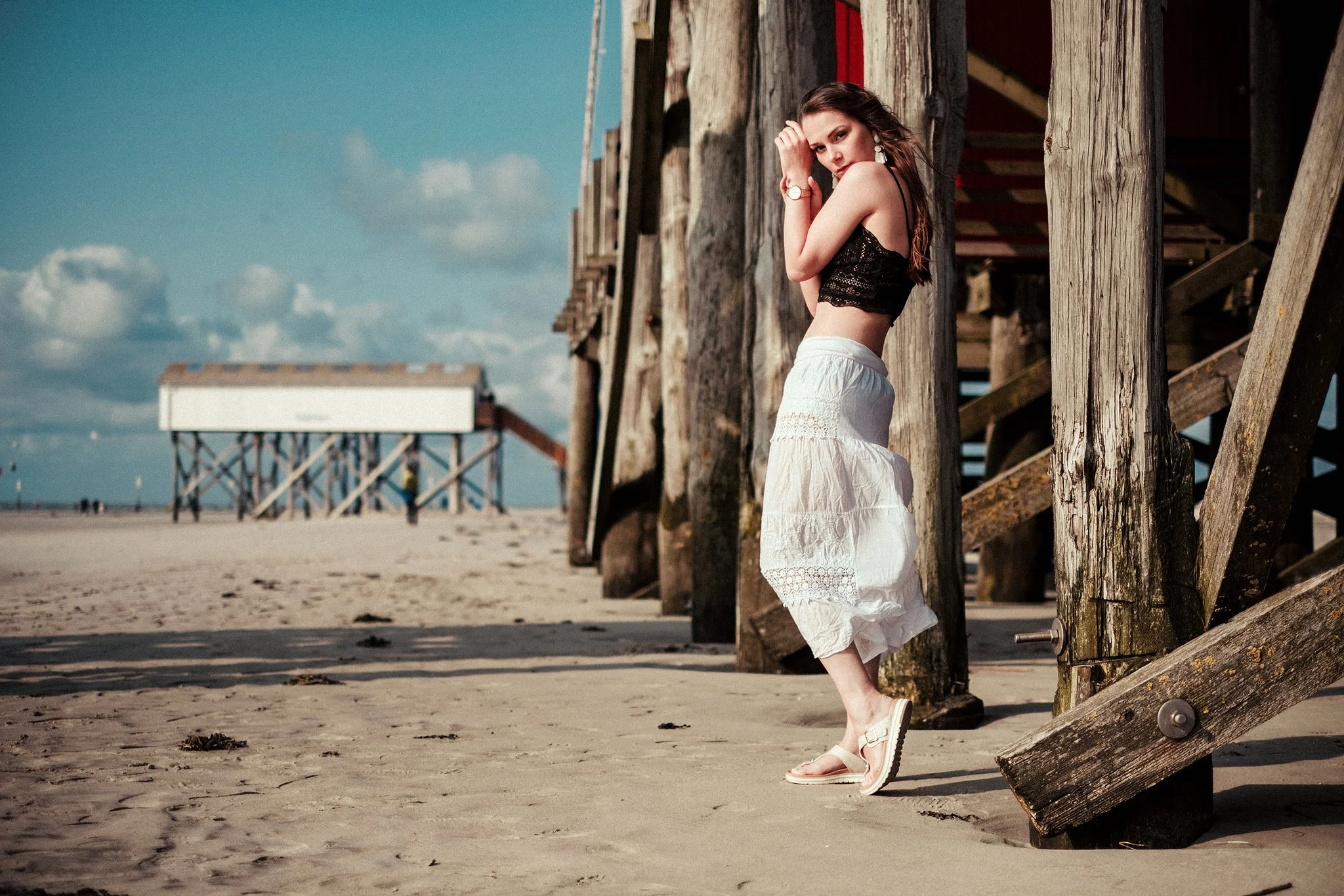 Frau in Sommerkleidung steht am Strand neben hölzernem Pfahlbau.