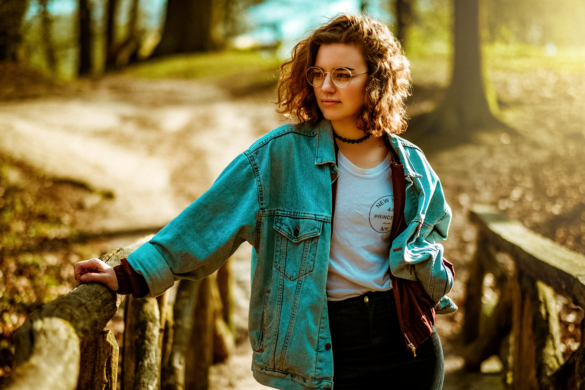 Frau mit lockigem Haar und Brille steht auf einem hölzernen Weg im Wald, trägt eine Jeansjacke und ein weißes T-Shirt.