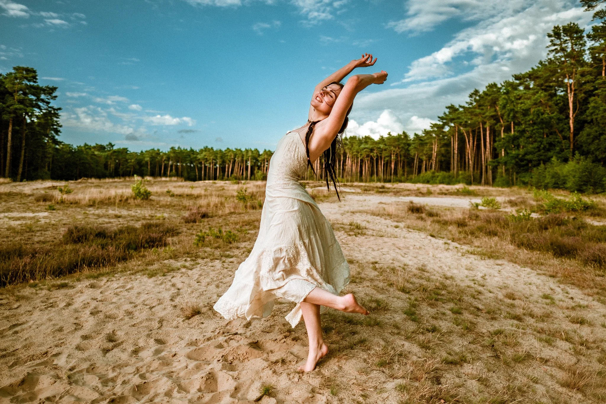 Eine Frau in einem weißen Kleid tanzt barfuß in einer sandigen Wiese vor einem Hintergrund aus Bäumen und blauem Himmel.