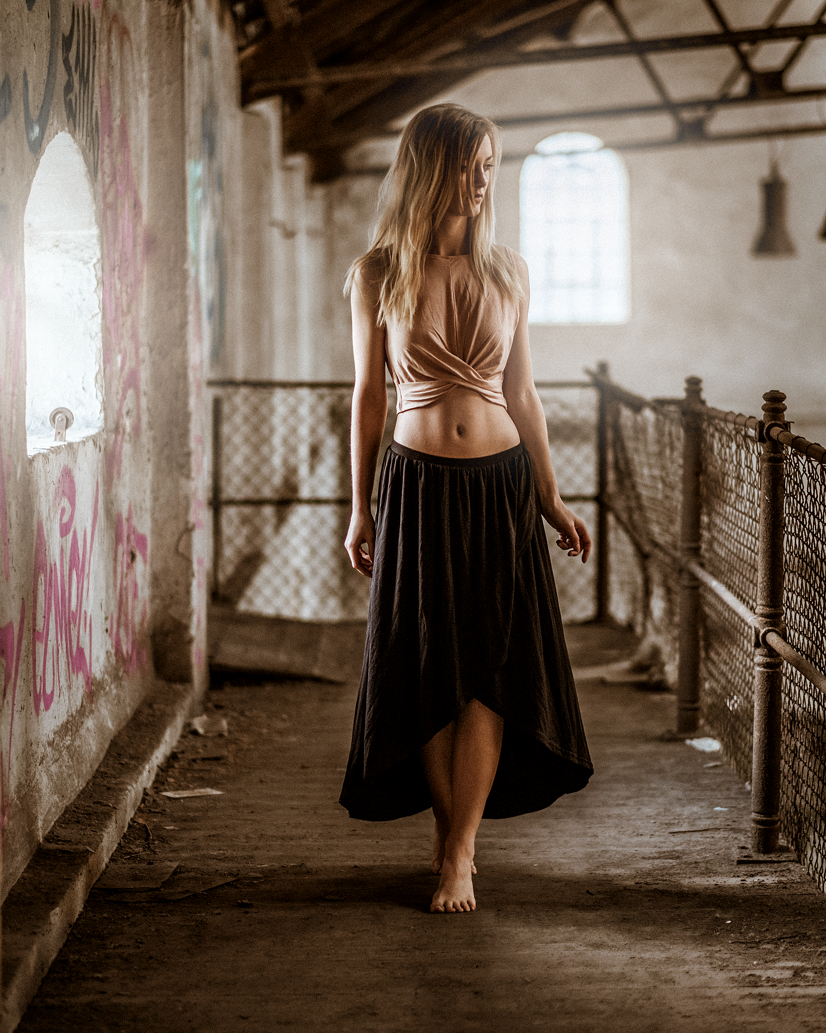 Frau in einem alten, verlassenen Gebäude, barfuß, trägt einen langen Rock und ein Oberteil, steht nah an einer Graffiti-bedeckten Wand.