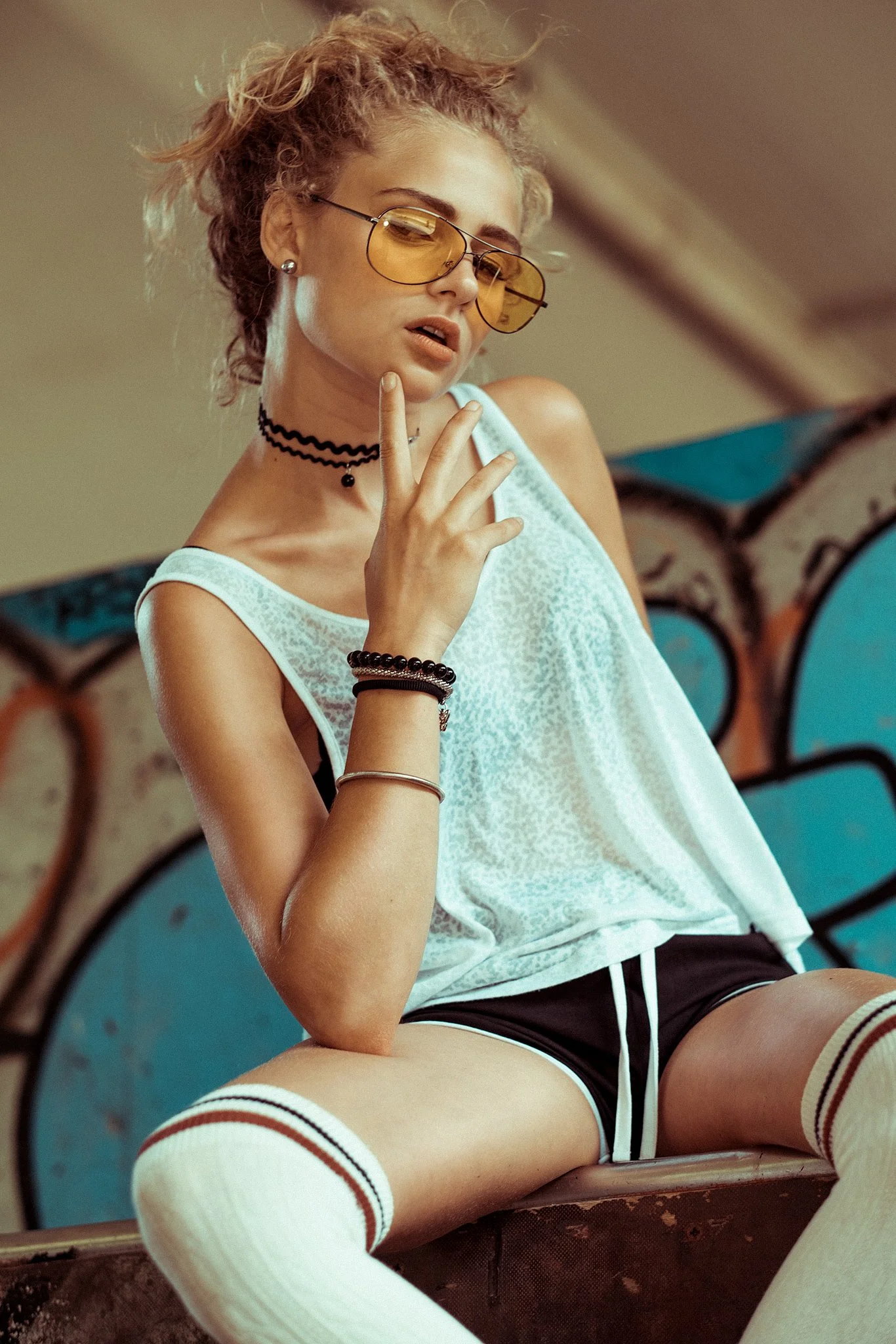 Person mit gelber Sonnenbrille, weißem Tanktop und Kniestrümpfen vor Graffiti.