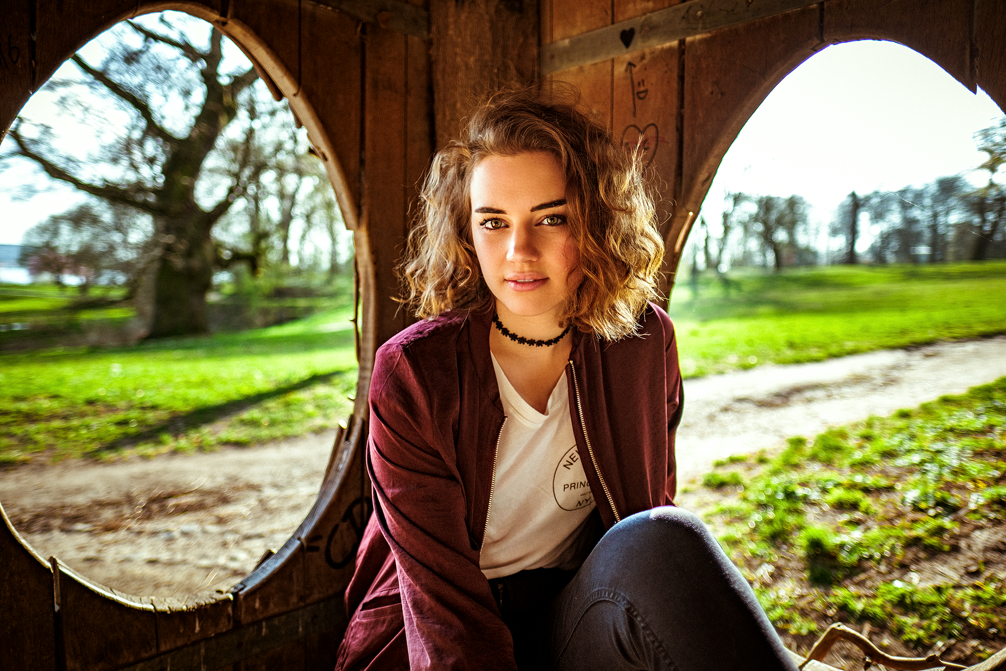 Junge Frau sitzt in einem hölzernen Pavillon mit ovalen Fenstern, umgeben von einem sonnigen Parkhintergrund.