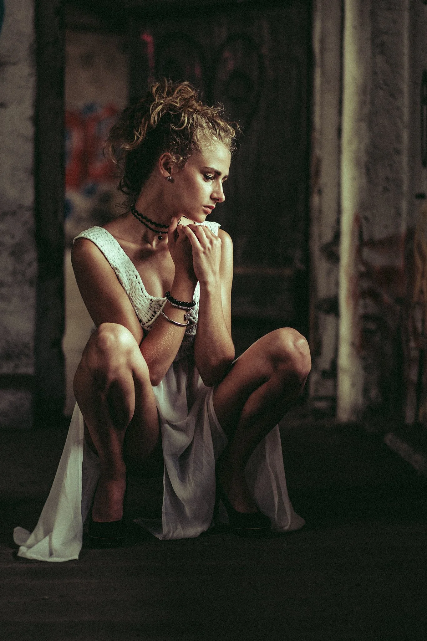 Frau in elegantem weißen Kleid hockt in einem dunklen Raum, umgeben von Graffiti.