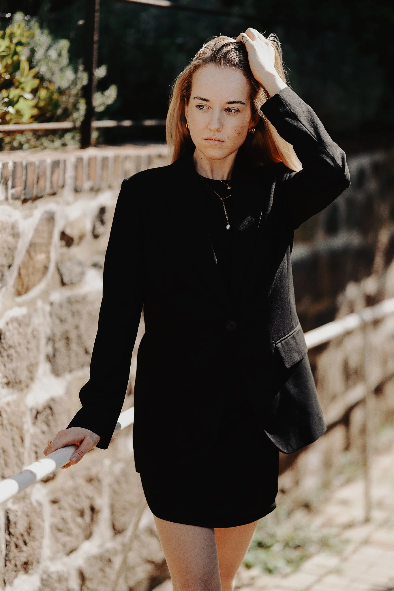 Frau in schwarzem Blazer und Rock an einer Steinmauer.