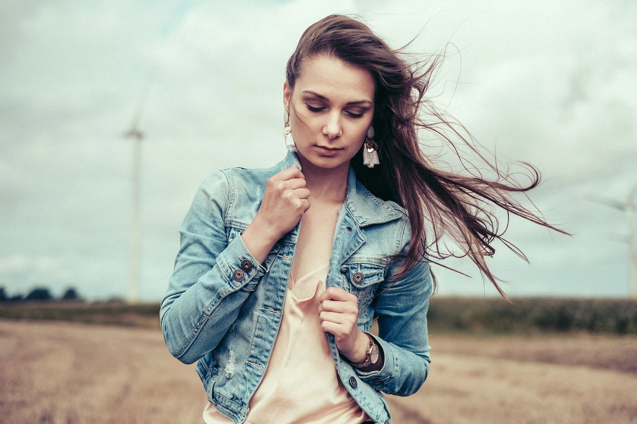 Frau im Jeansjacke steht auf einem Feld, ihr Haar weht im Wind. Im Hintergrund sind Windräder sichtbar.
