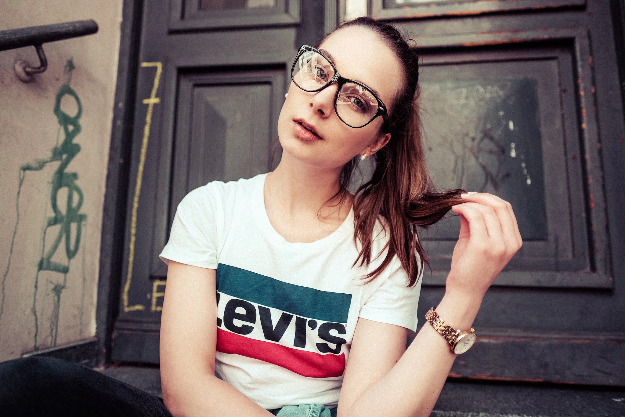 Junge Frau mit Brille und weißem Levi's T-Shirt sitzt auf einer Treppe vor einer Tür. Sie hält eine Haarsträhne mit der Hand und trägt eine Armbanduhr.