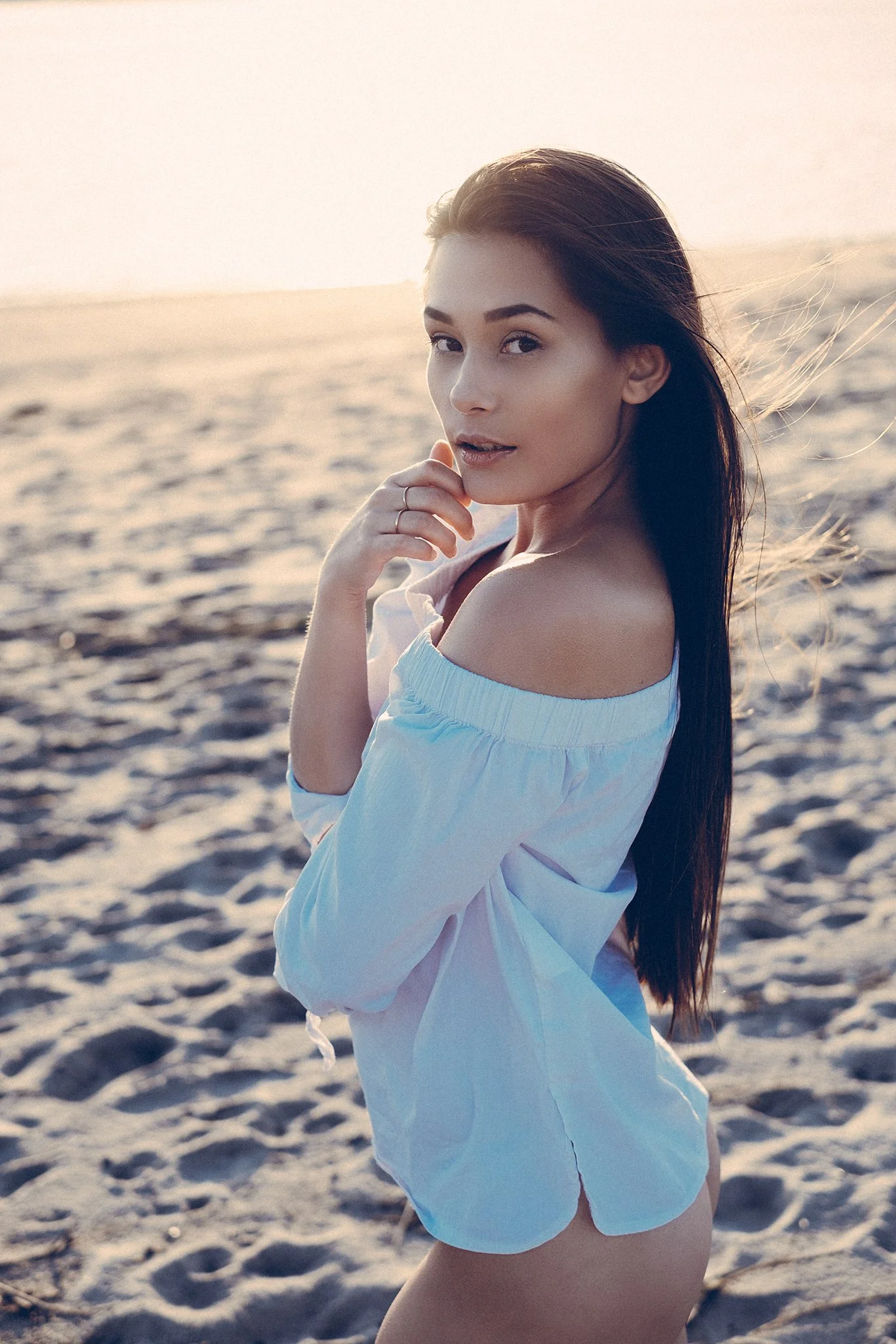 Eine Frau mit langen Haaren und einer weißen Bluse steht am Strand und blickt über die Schulter in die Kamera.
