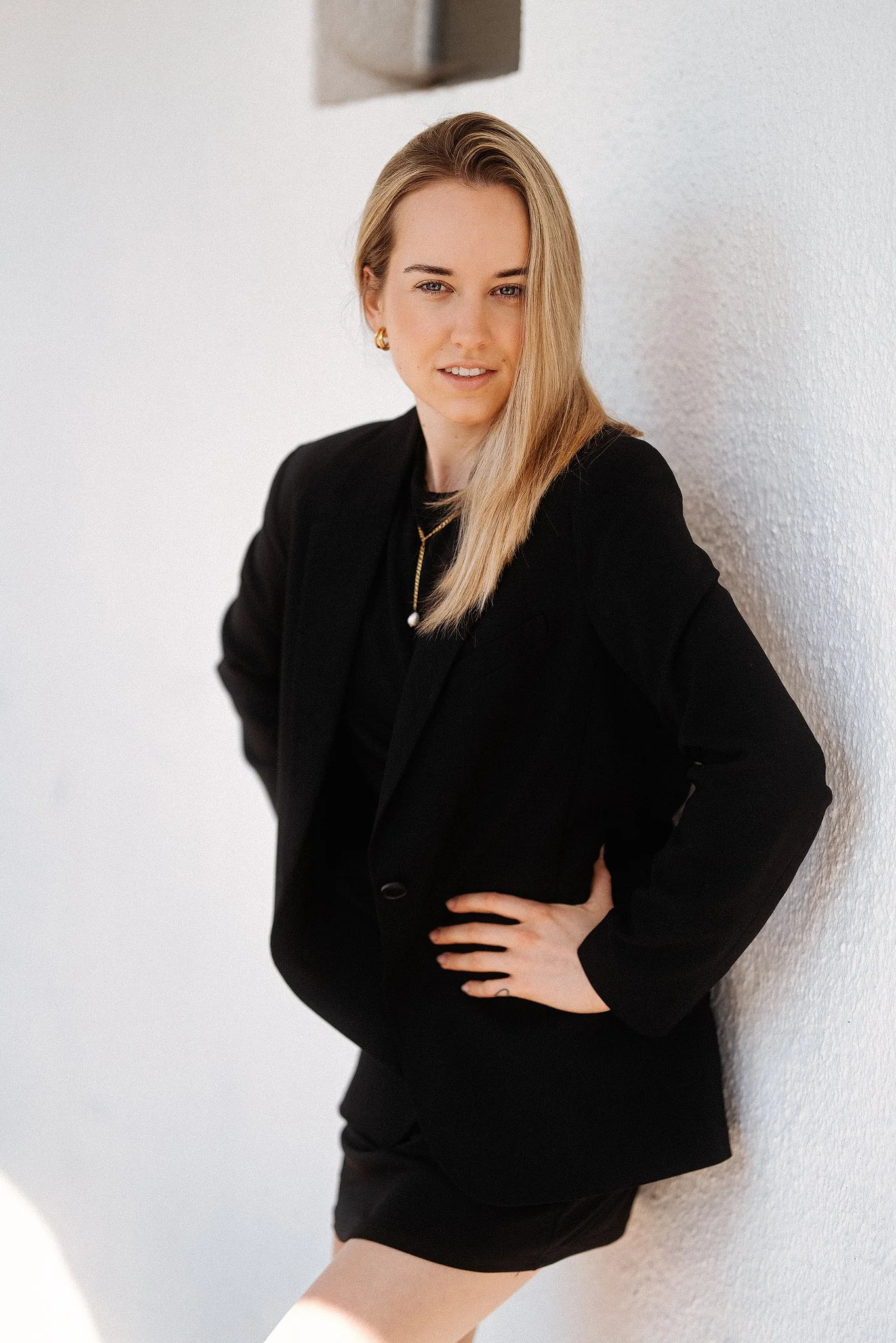 Frau in schwarzem Blazer, posiert an einer weißen Wand.
