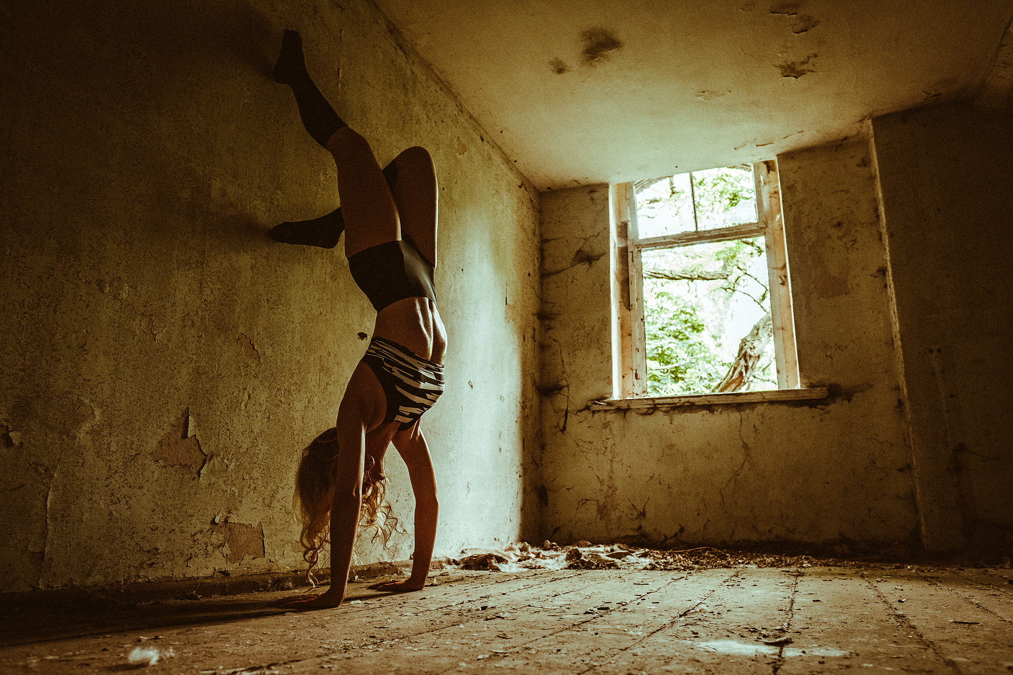 Person macht Handstand in einem verlassenen Raum mit abblätternden Wänden, neben einem Fenster mit Blick auf Bäume.
