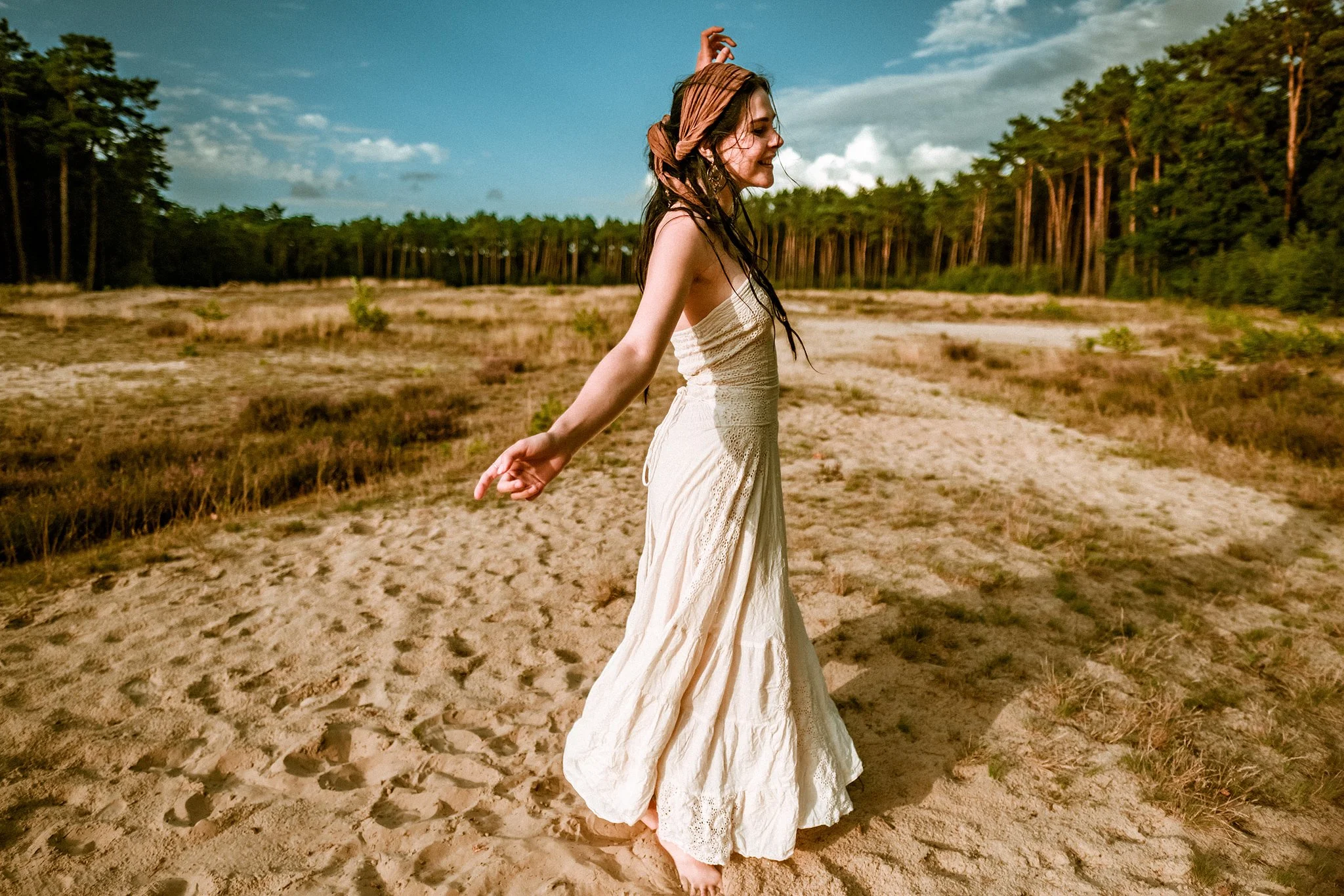 Frau in einem langen weißen Kleid steht barfuß auf einem sandigen Feld mit Bäumen im Hintergrund.