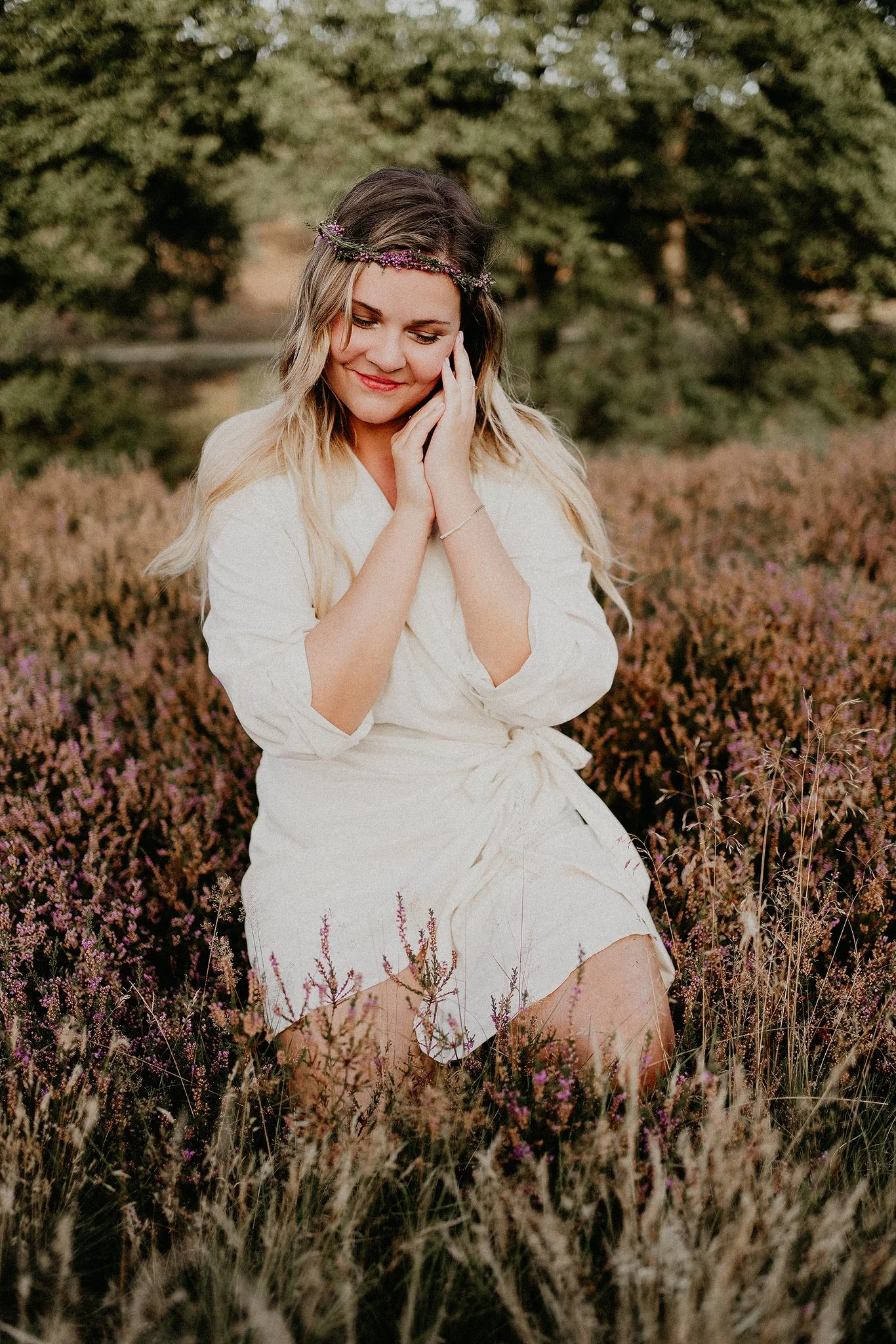 Frau in weißem Kleid sitzt in einer Heidewiese mit Blumenkranz im Haar, lächelnd, Hände an den Wangen, vor unscharfem Hintergrund mit Bäumen.