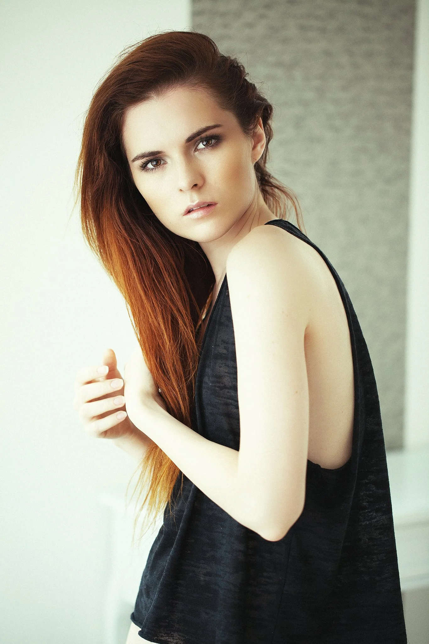 Porträt einer Frau mit langen, roten Haaren, die ein ärmelloses schwarzes Oberteil trägt; hell beleuchteter Hintergrund.