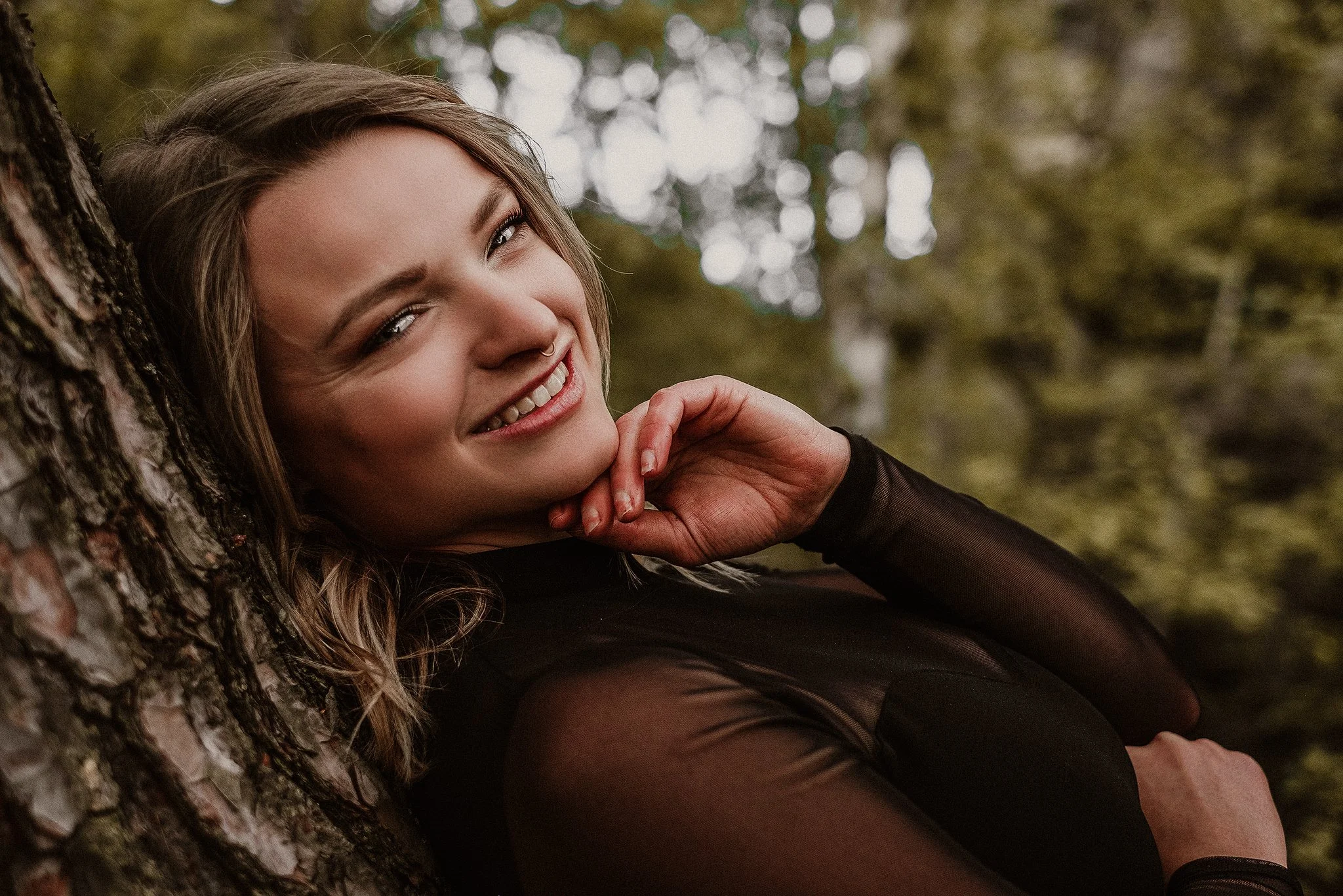 Lächelnde Frau lehnt sich an einen Baum, trägt schwarzes Oberteil, verschwommener Hintergrund aus Bäumen.