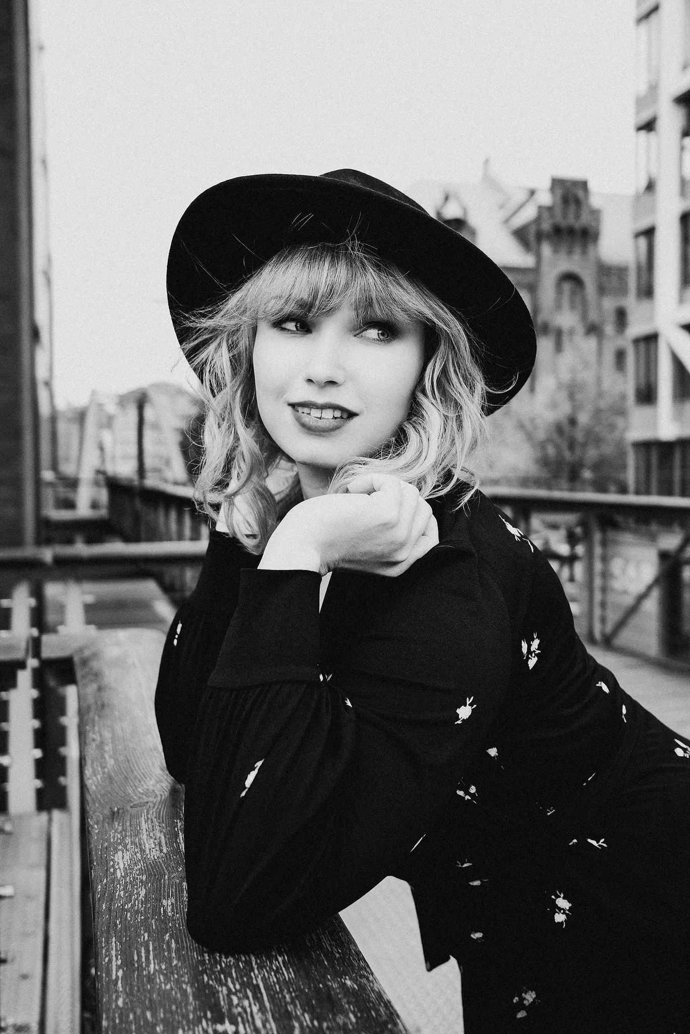 Schwarz-weiß-Foto einer Frau mit Hut, die lächelnd in die Ferne schaut. Sie lehnt sich auf einem Holzgeländer in einer städtischen Umgebung.