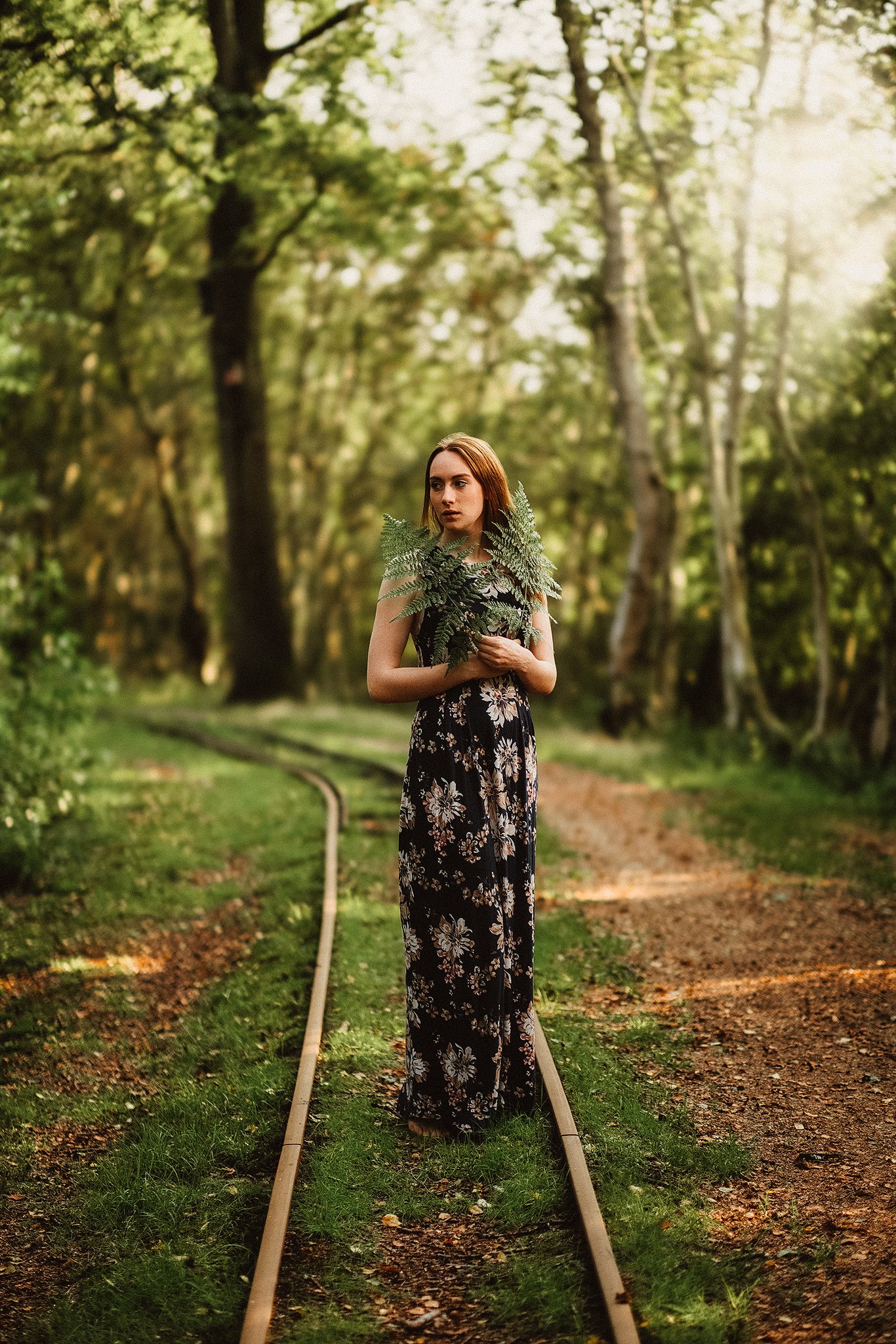 Eine Frau in einem langen, geblümten Kleid steht auf einer stillgelegten Eisenbahnschiene in einem Wald und hält Farnzweige.