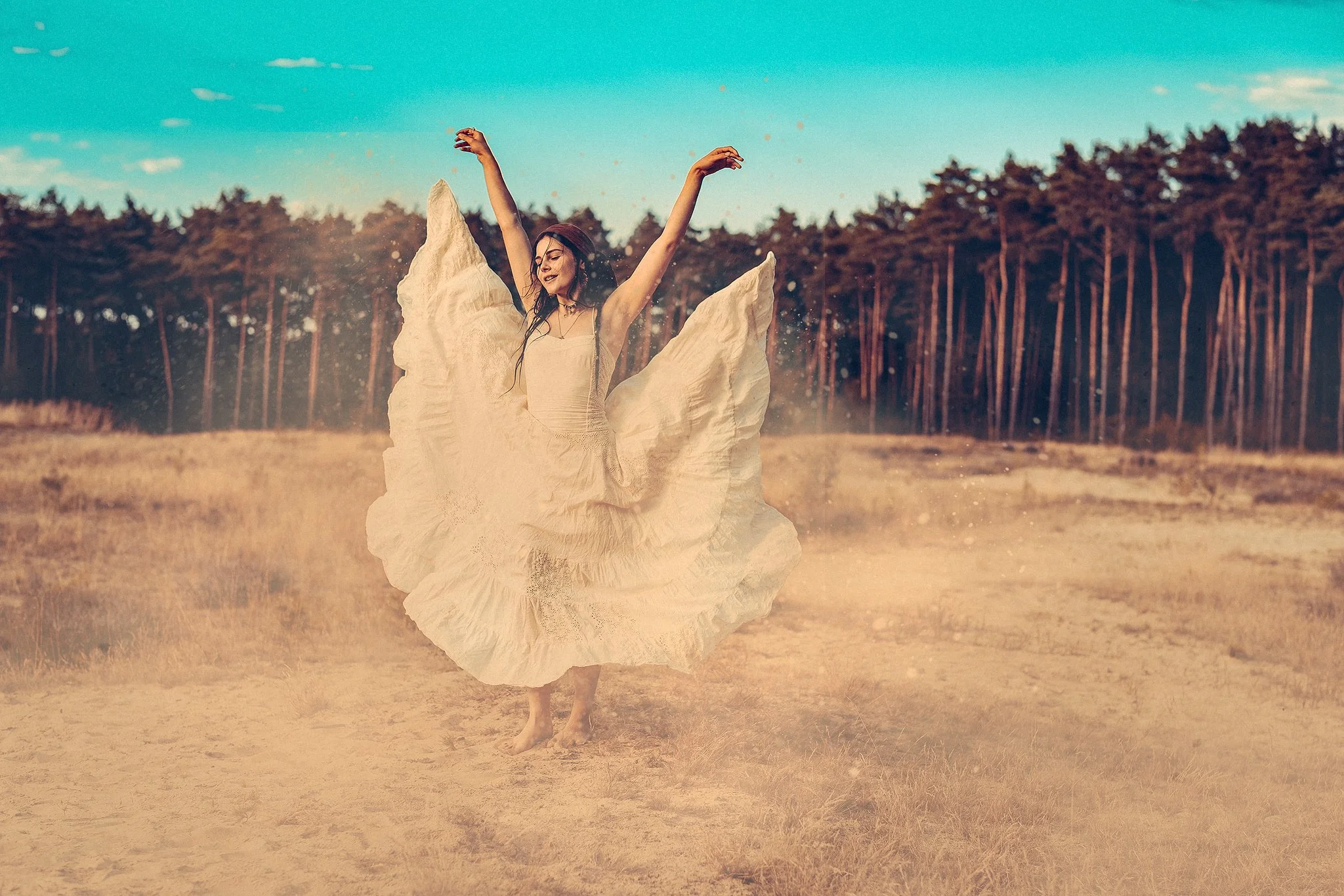 Frau in langem weißen Kleid tanzt freudig in einer sandigen Landschaft vor einem Wald.