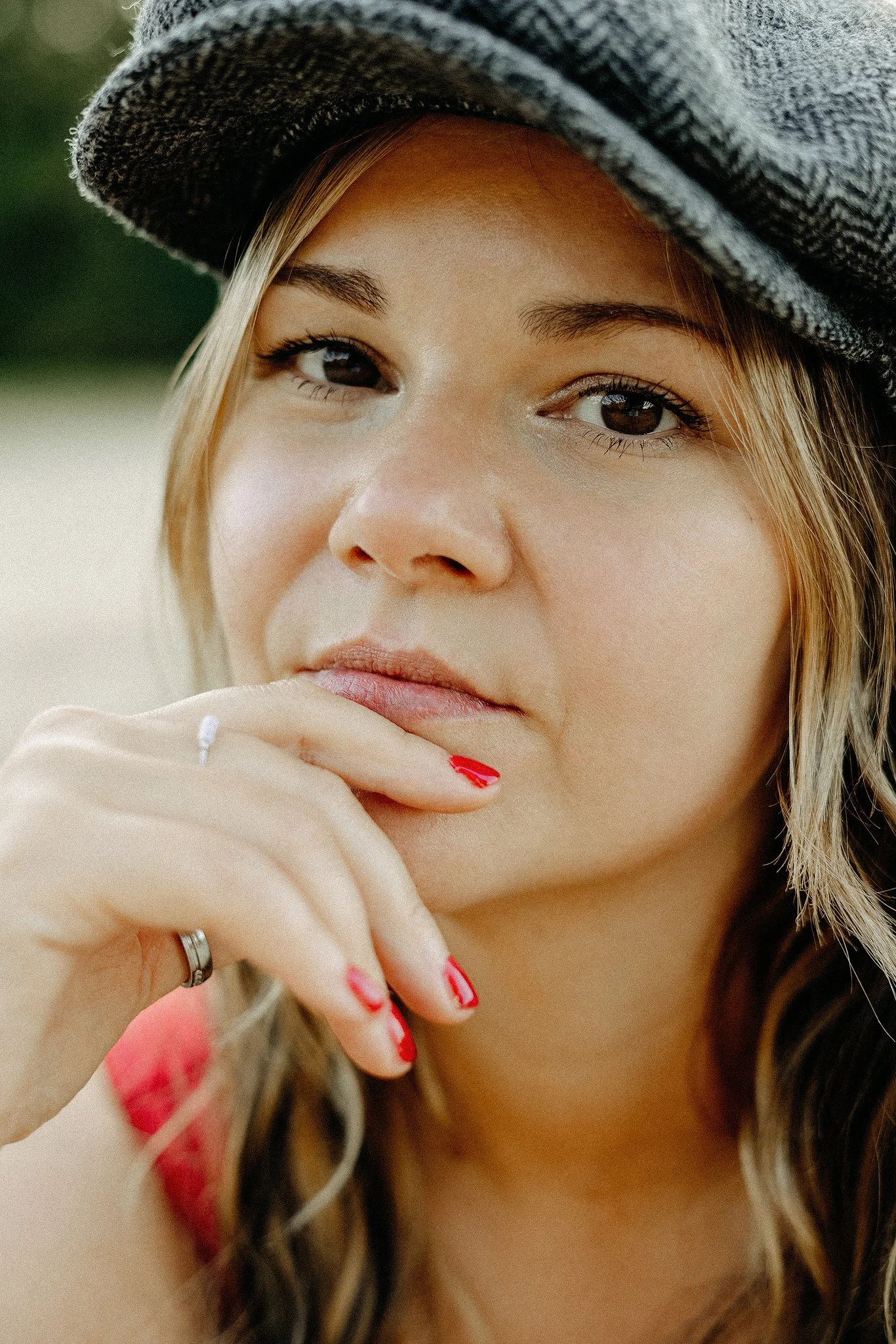 Nahaufnahme einer Frau mit grauer Mütze und roten Fingernägeln, die nachdenklich aussieht und ihre Hand ans Kinn legt.