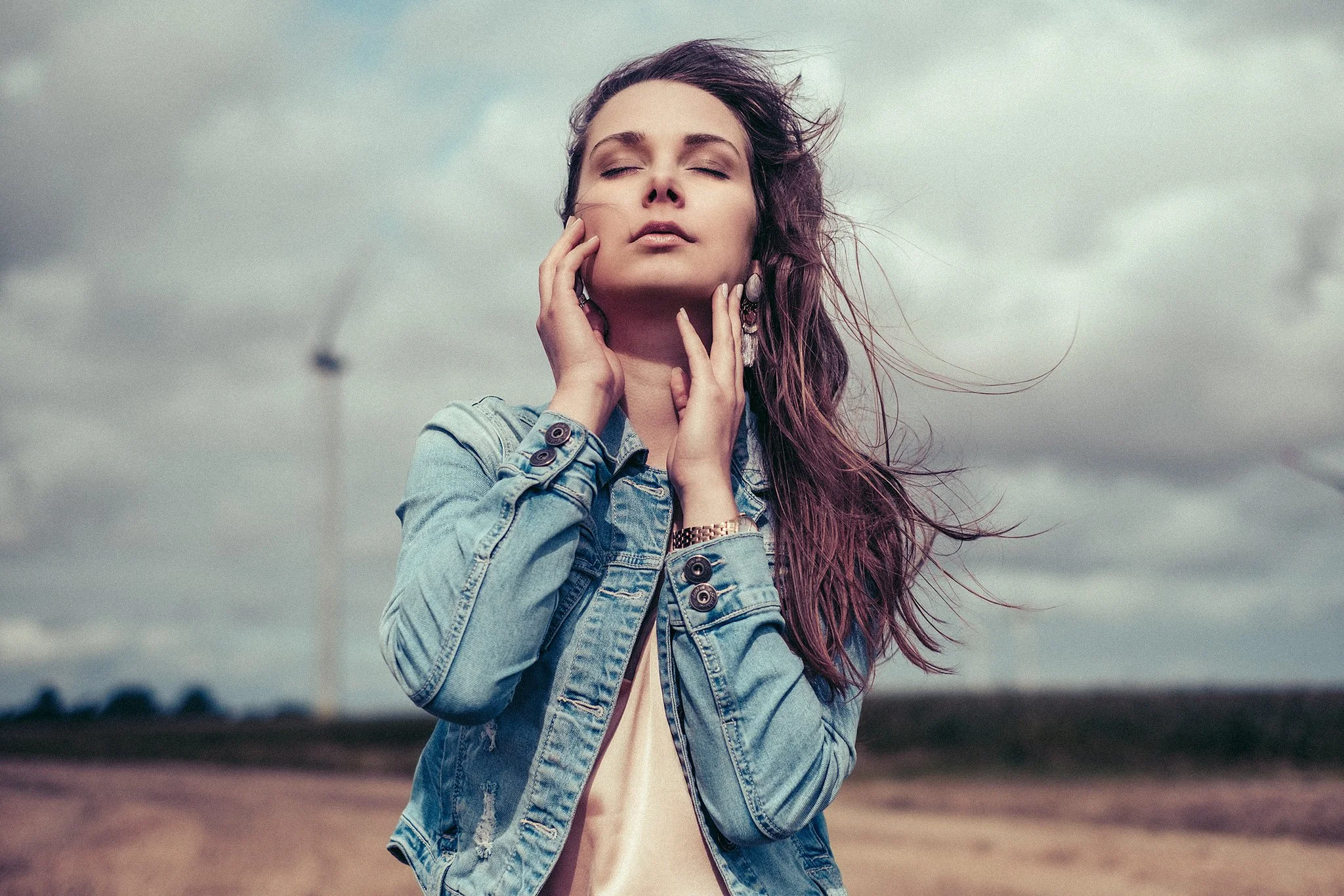 Frau mit geschlossenen Augen in Jeansjacke vor bewölktem Himmel in einer Landschaft.