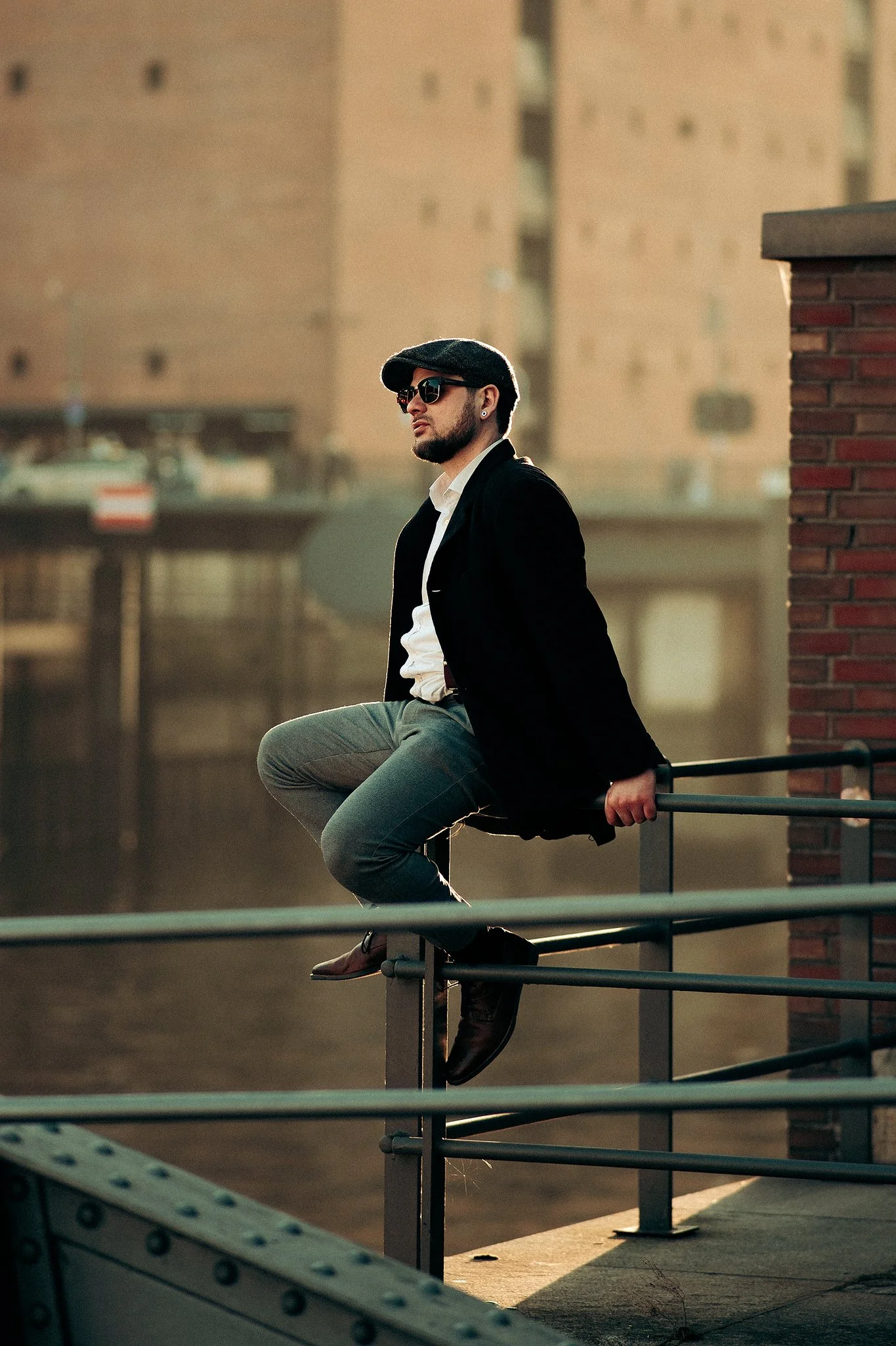 Mann mit Bart sitzt auf einem Geländer, trägt Mütze und Sonnenbrille, vor unscharfem Stadt-Hintergrund in warmem Licht.