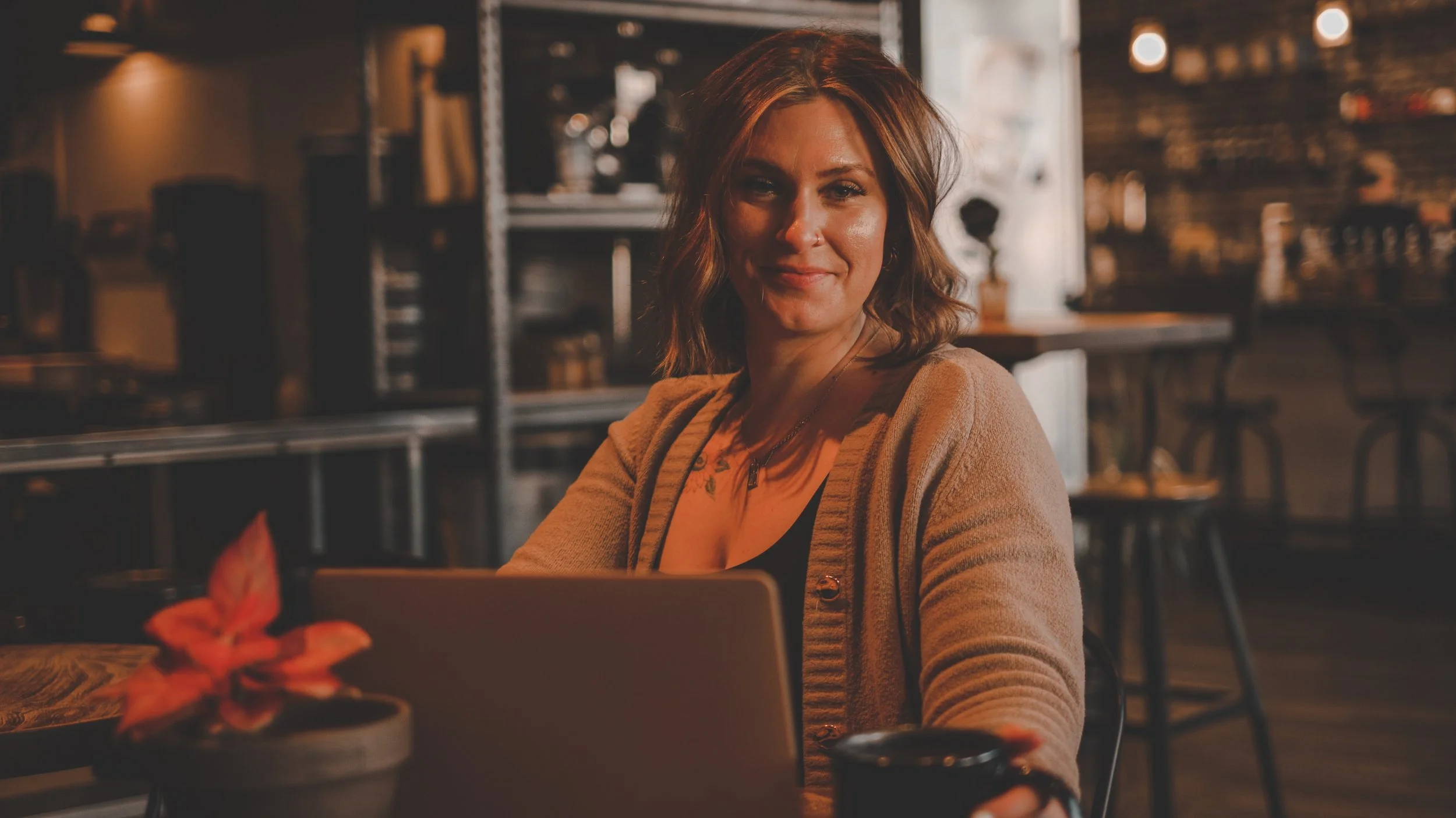 A woman with shoulder-length brown hair smiling and sitting in a cozy, warmly lit cafe or coffee shop, with a laptop in front of her, a small potted plant with red and pink leaves nearby, and a blurred background of shelves and tables.