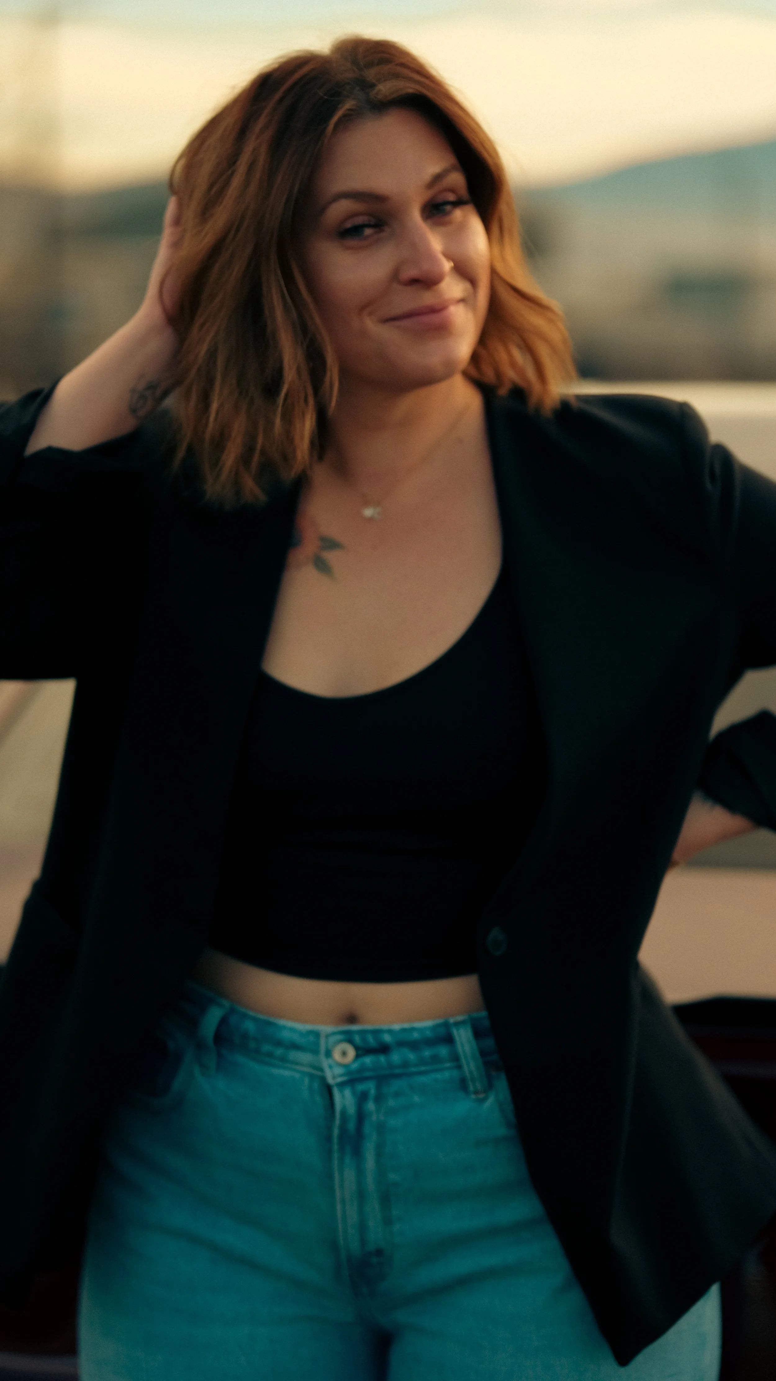 A woman with shoulder-length, reddish-brown hair, wearing a black crop top, black blazer, and light blue jeans, standing outdoors with her hand behind her head and a slight smile.