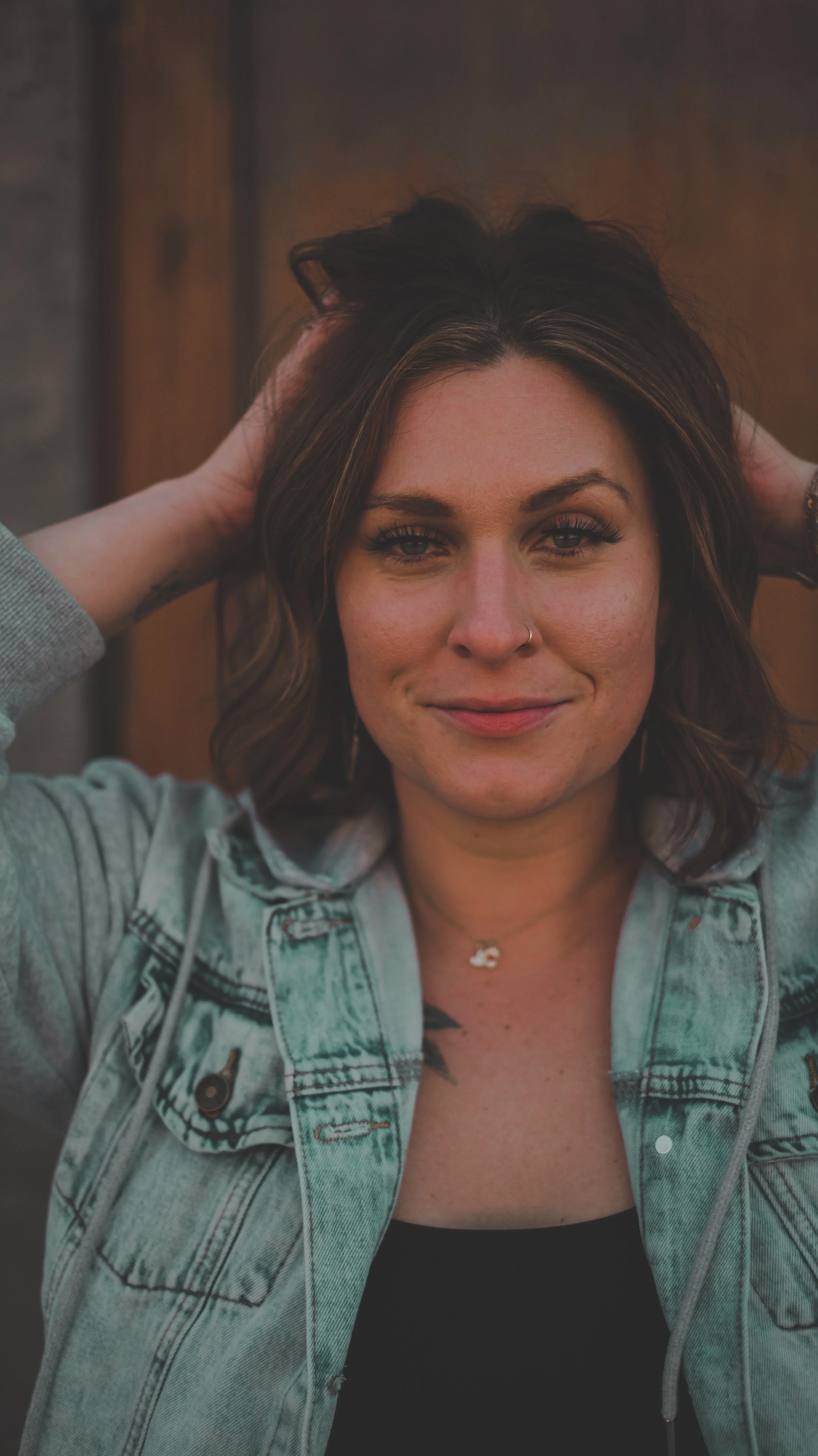 A woman with shoulder-length brown hair and a nose ring, smiling and holding her hands behind her head, wearing a denim jacket over a black top.