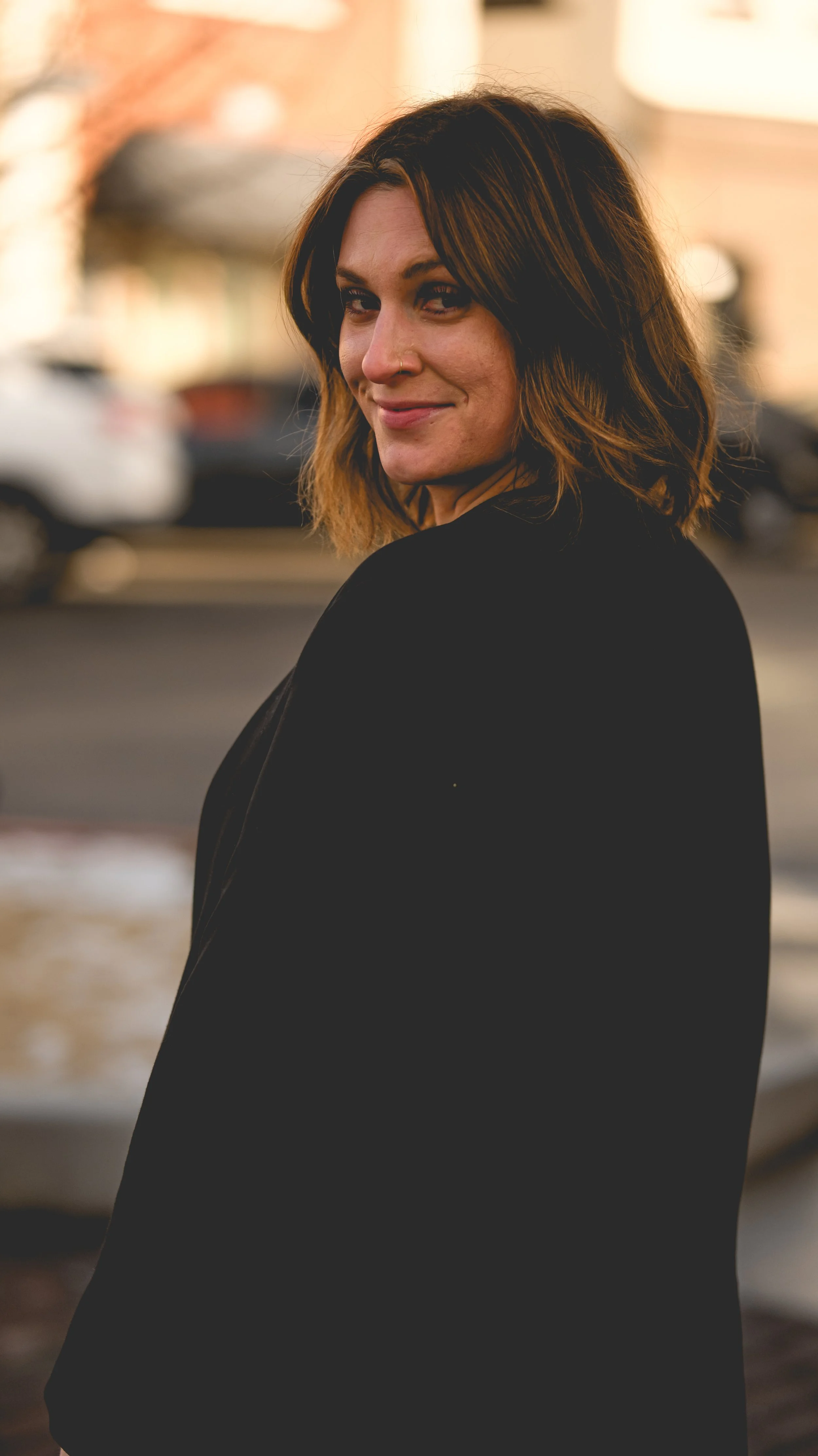A woman with shoulder-length brown hair, smiling, wearing a black jacket, standing outdoors with a blurred city street background.