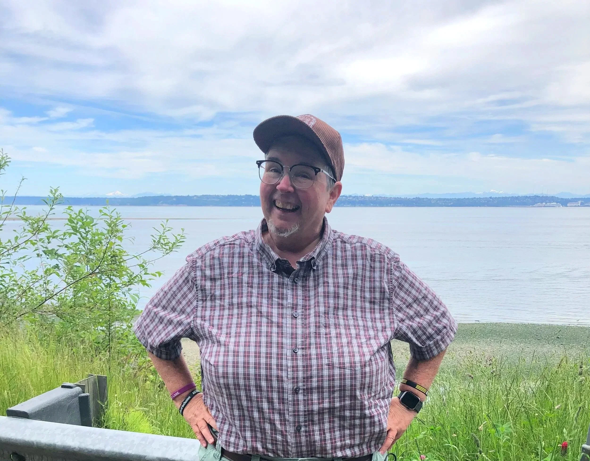 image of a human wearing a brown corduroy hat and wearing glasses and a checkered button down shirt, standing in front of a cloudy sky, a cityscape and water.